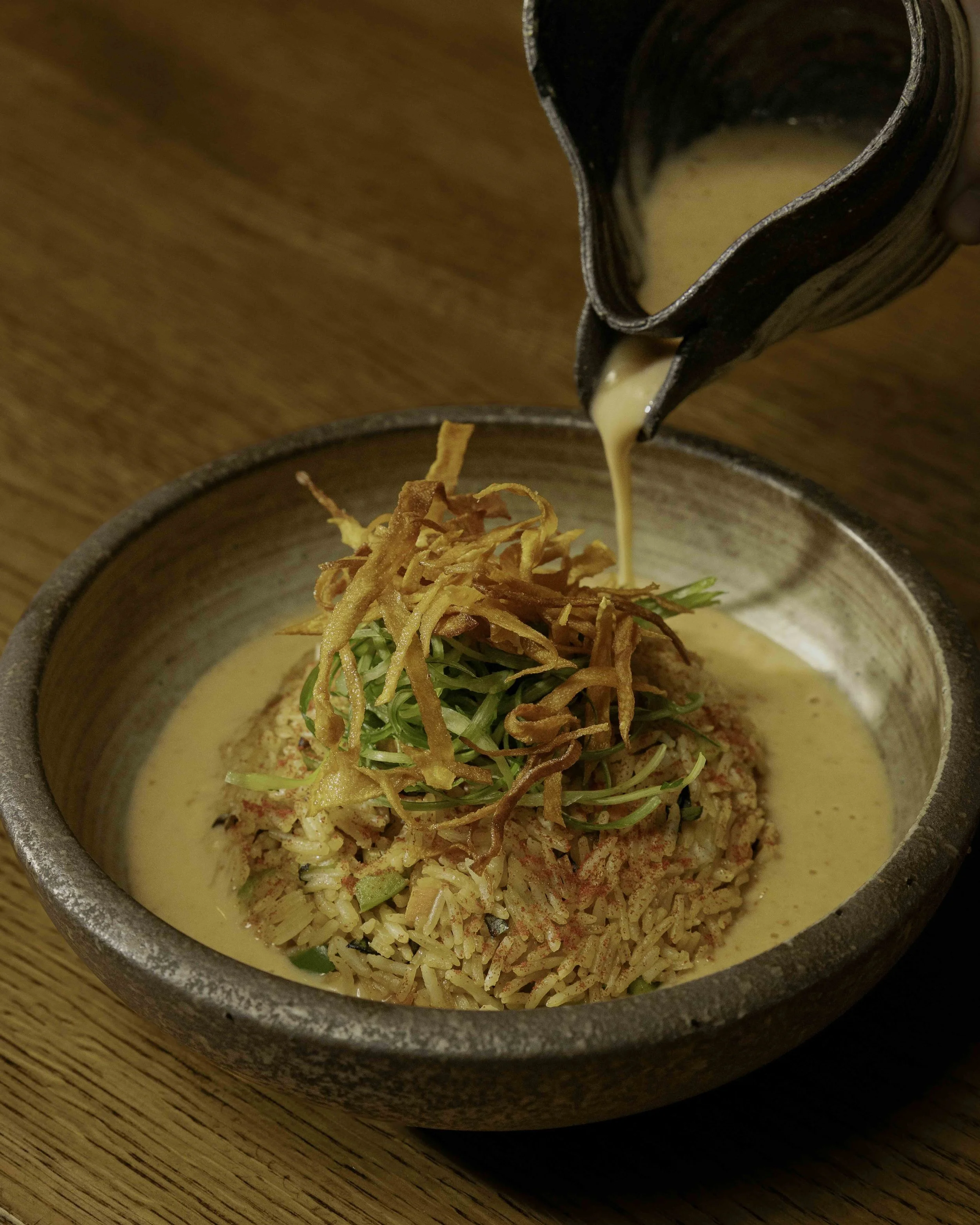 A bowl of ramen noodles topped with fried onions and green garnish, as broth is poured over from a pitcher.
