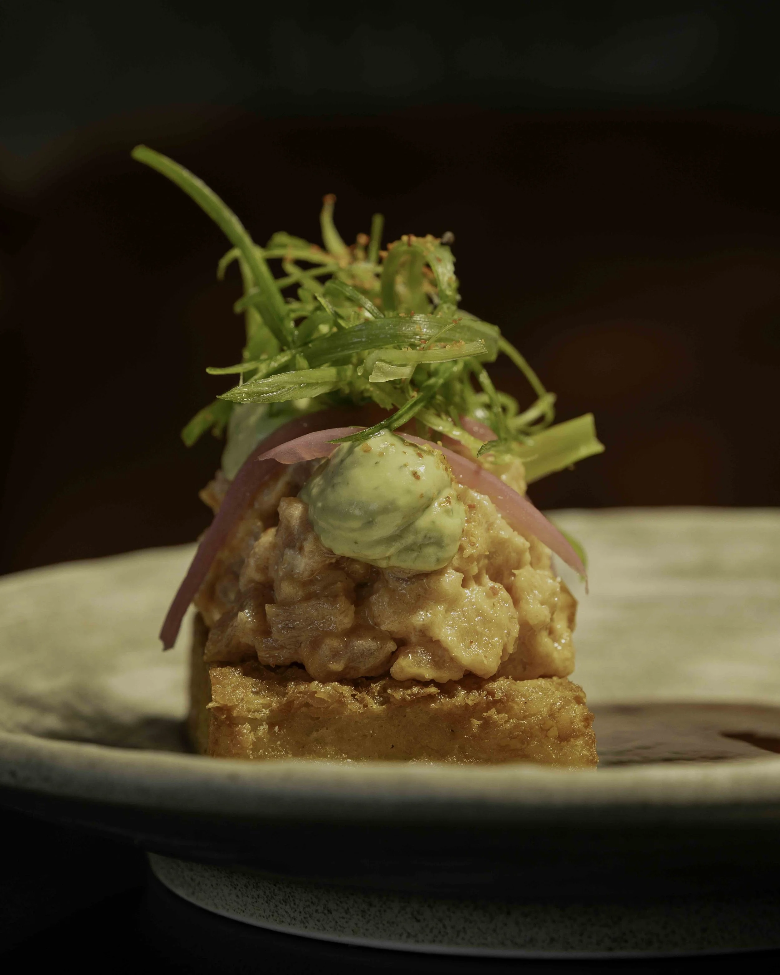 Close-up of a plated dish with a fried item topped with creamy sauce, pink onions, green herbs, and garnished with fresh greens.