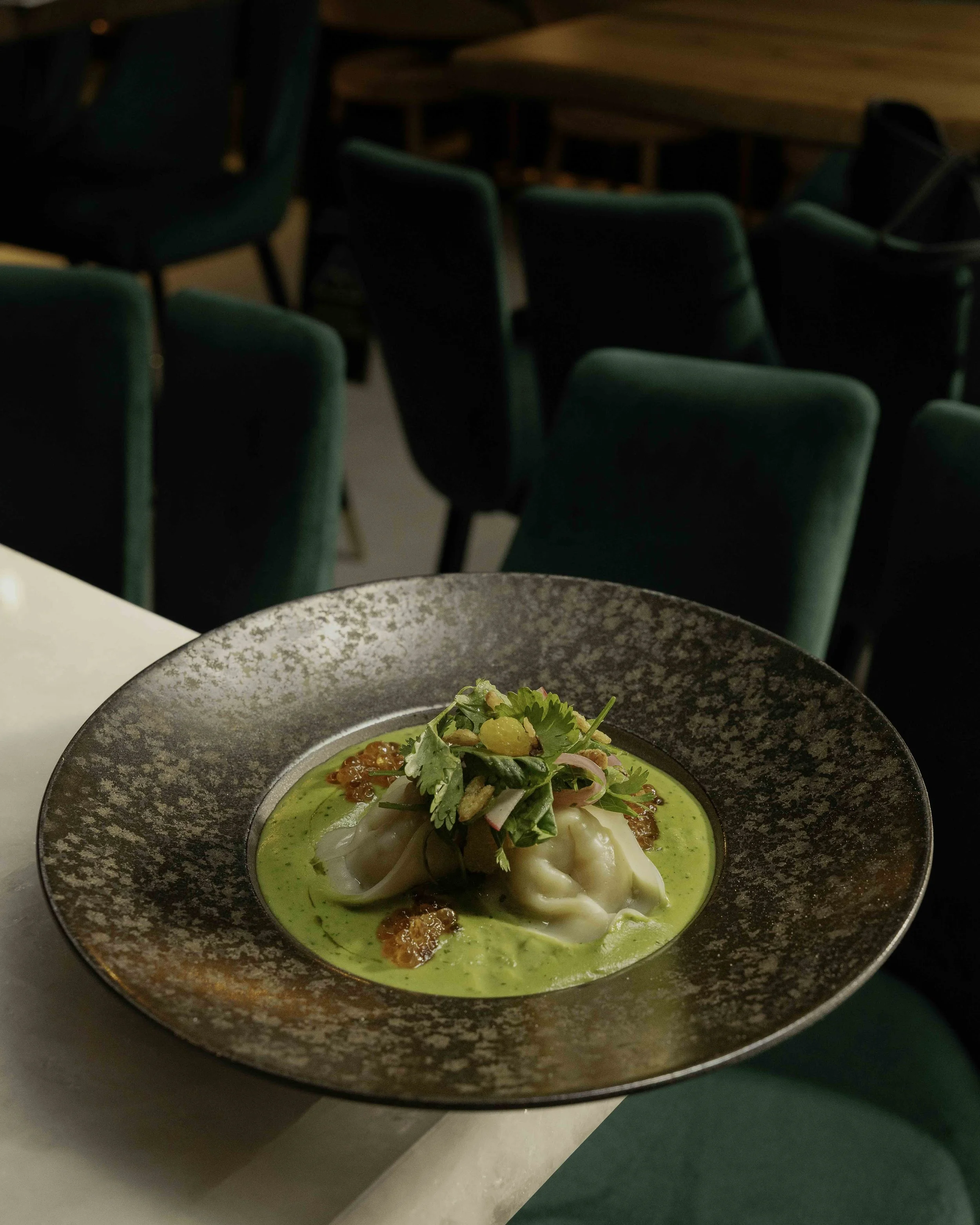 A plate of Asian dumplings served in a green sauce, garnished with herbs, scallions, and roe, on a textured dark plate in a restaurant setting with green velvet chairs in the background.