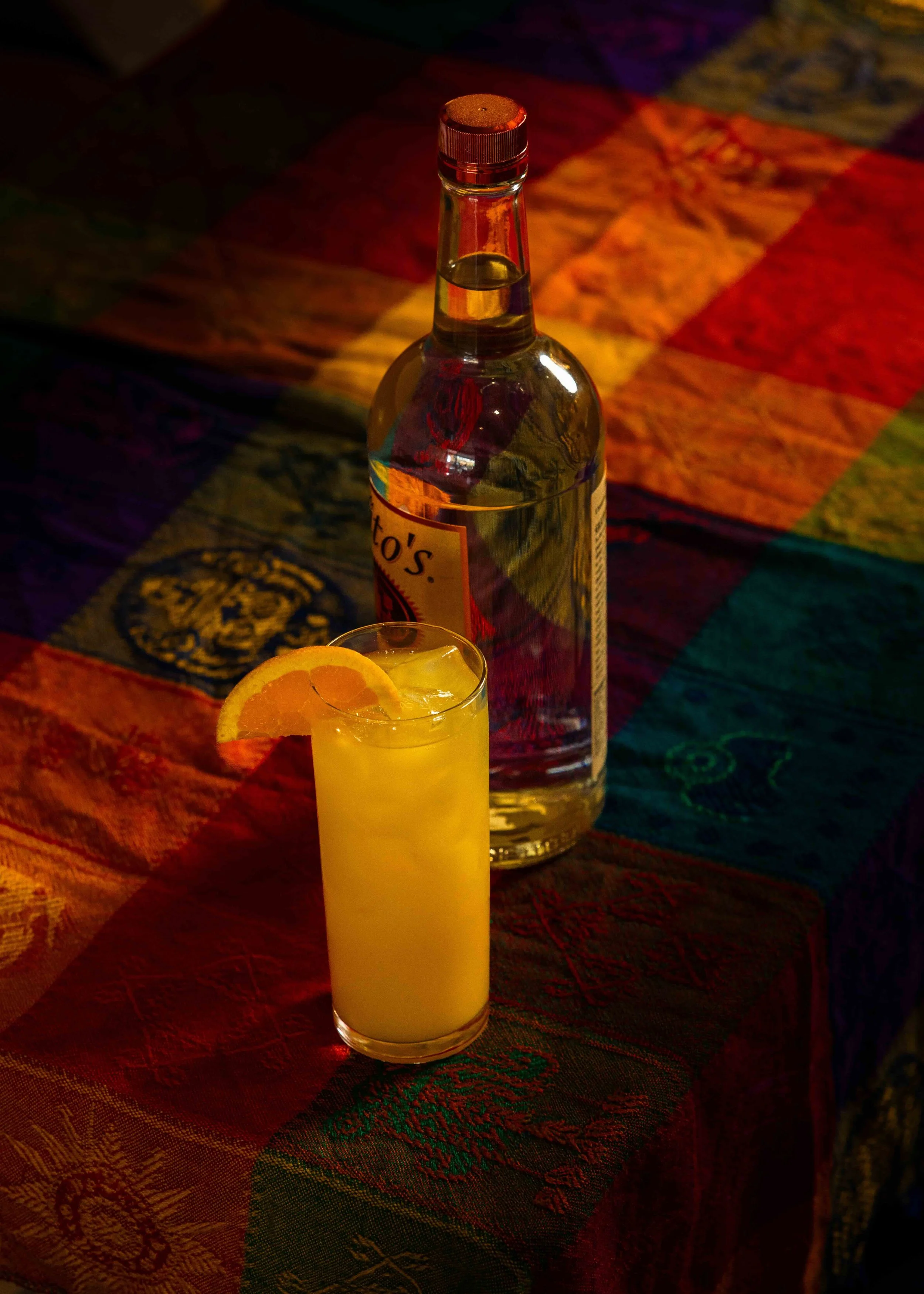 A bottle of alcohol and a tall glass of a yellow cocktail with a lemon slice on the rim are on a colorful, patterned tablecloth.