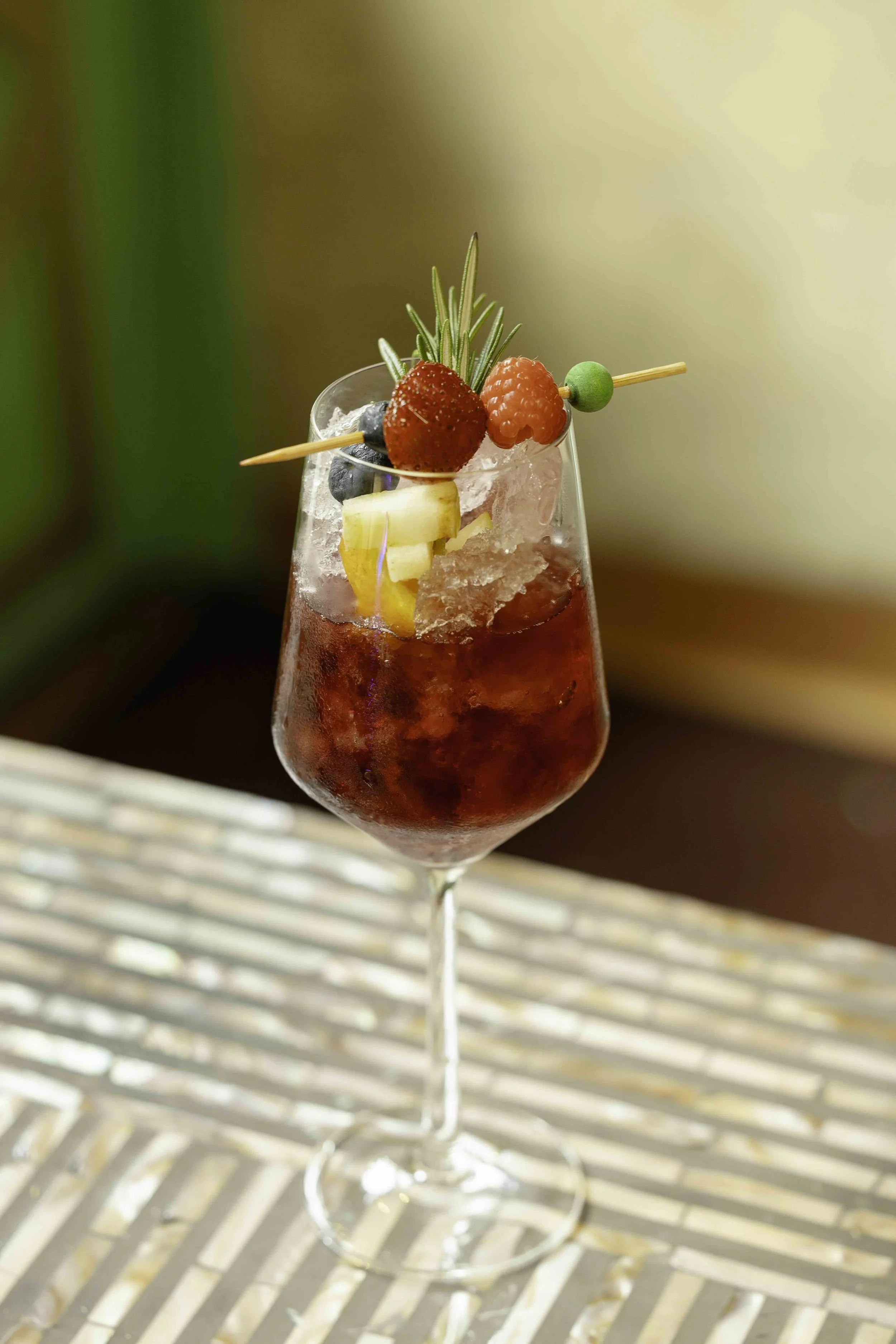 A tall glass of mixed berry cocktail with ice, garnished with fresh strawberries, raspberries, blueberries, a small piece of pineapple, and a sprig of rosemary.