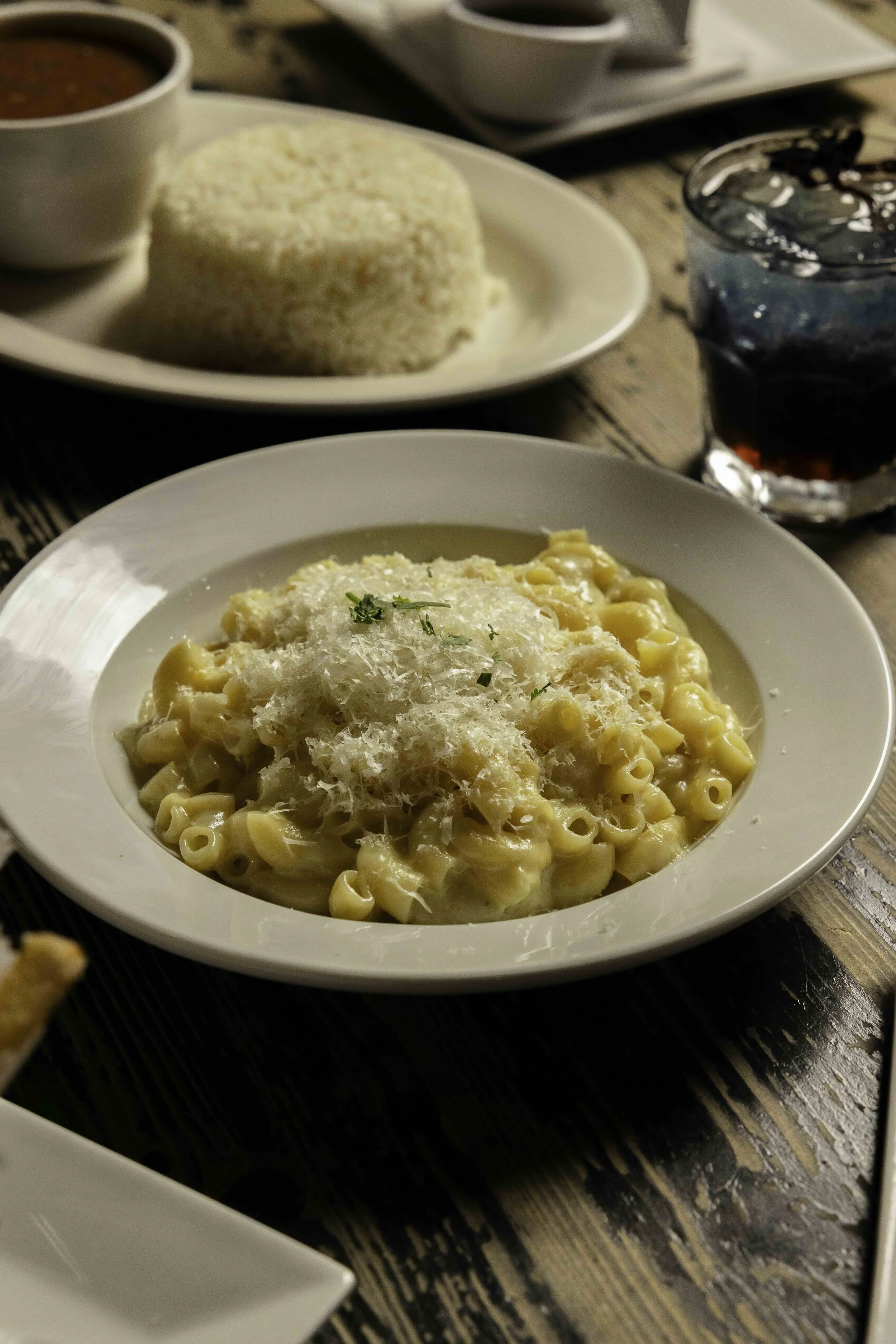 Creamy macaroni and cheese topped with grated cheese and herbs on a white plate, with a glass of soda in the background.