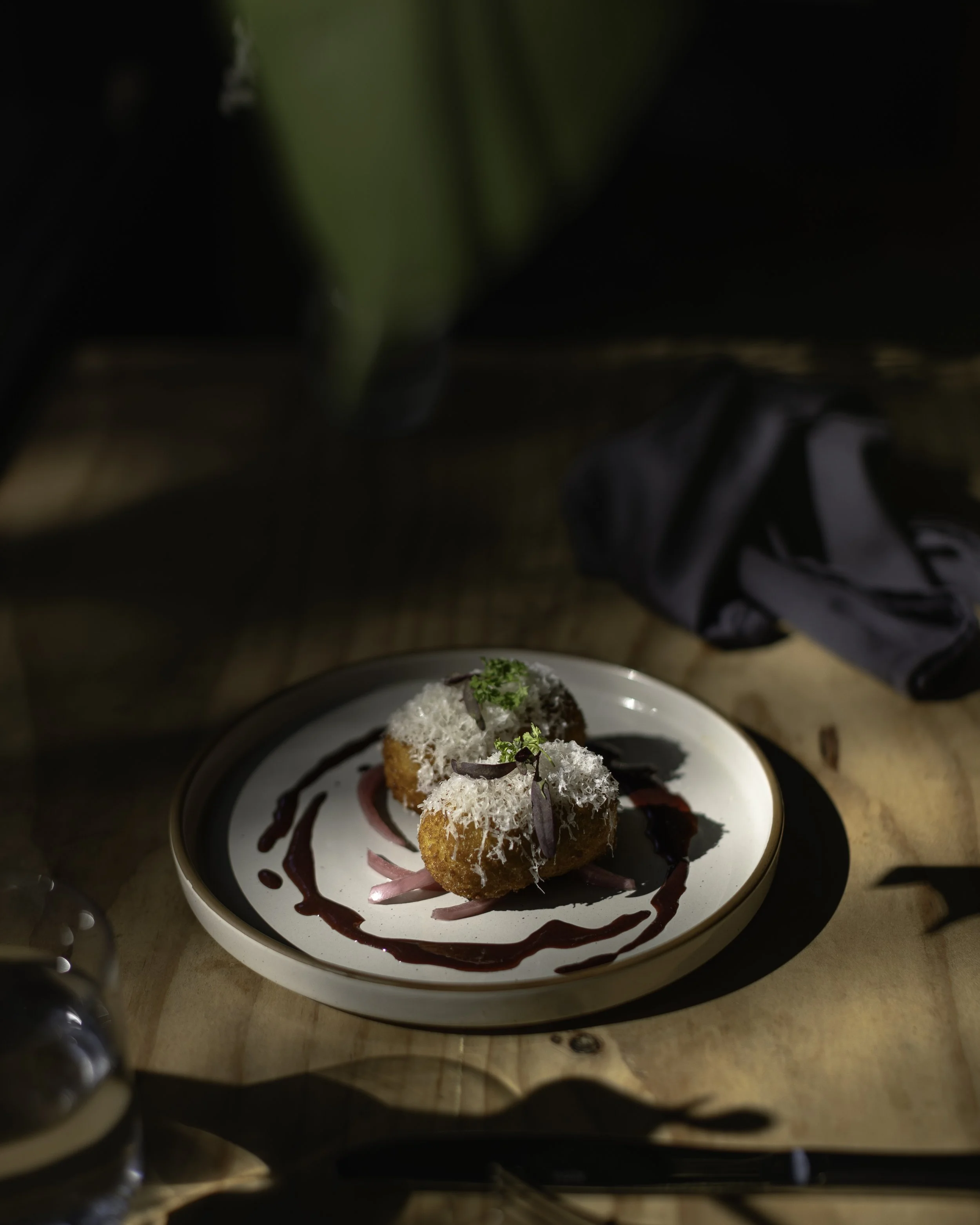 Two fried appetizers topped with shredded cheese, garnished with herbs, served with a dark sauce drizzle on a white plate, on a wooden table.
