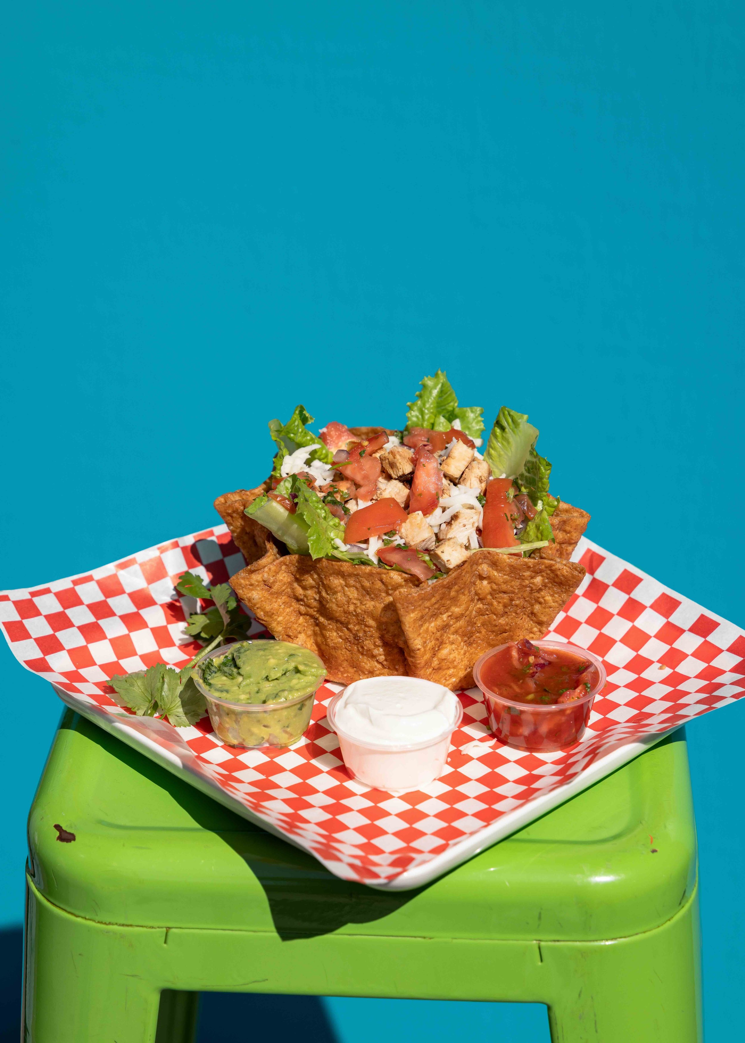 Tacos with lettuce, tomatoes, shredded cheese, and grilled chicken, served in a crispy shell, with guacamole, sour cream, and salsa on the side, on a red checkered paper on a green plastic table against a blue background.