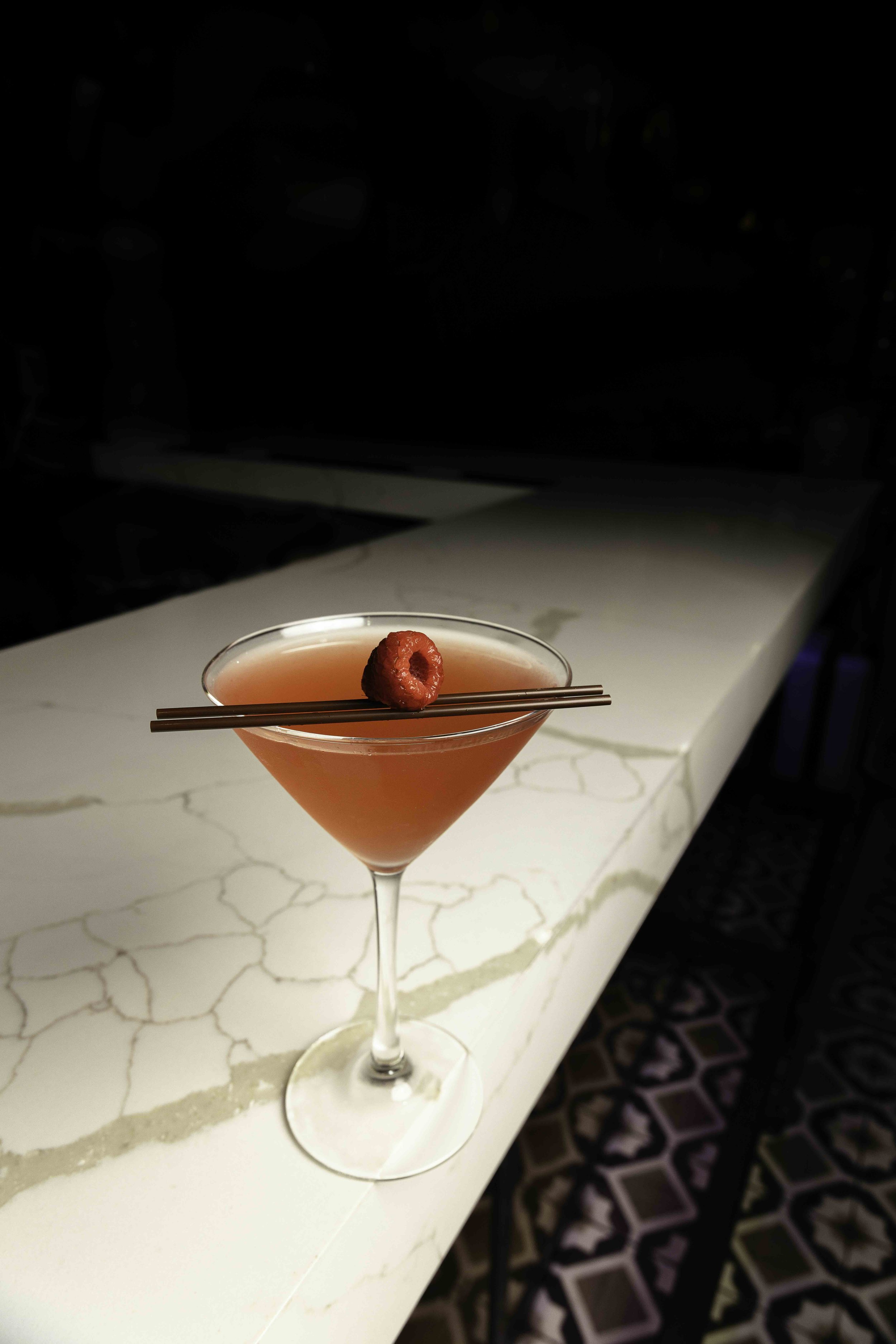A pink cocktail garnished with a raspberry and two chocolate sticks, placed on a white marble surface.