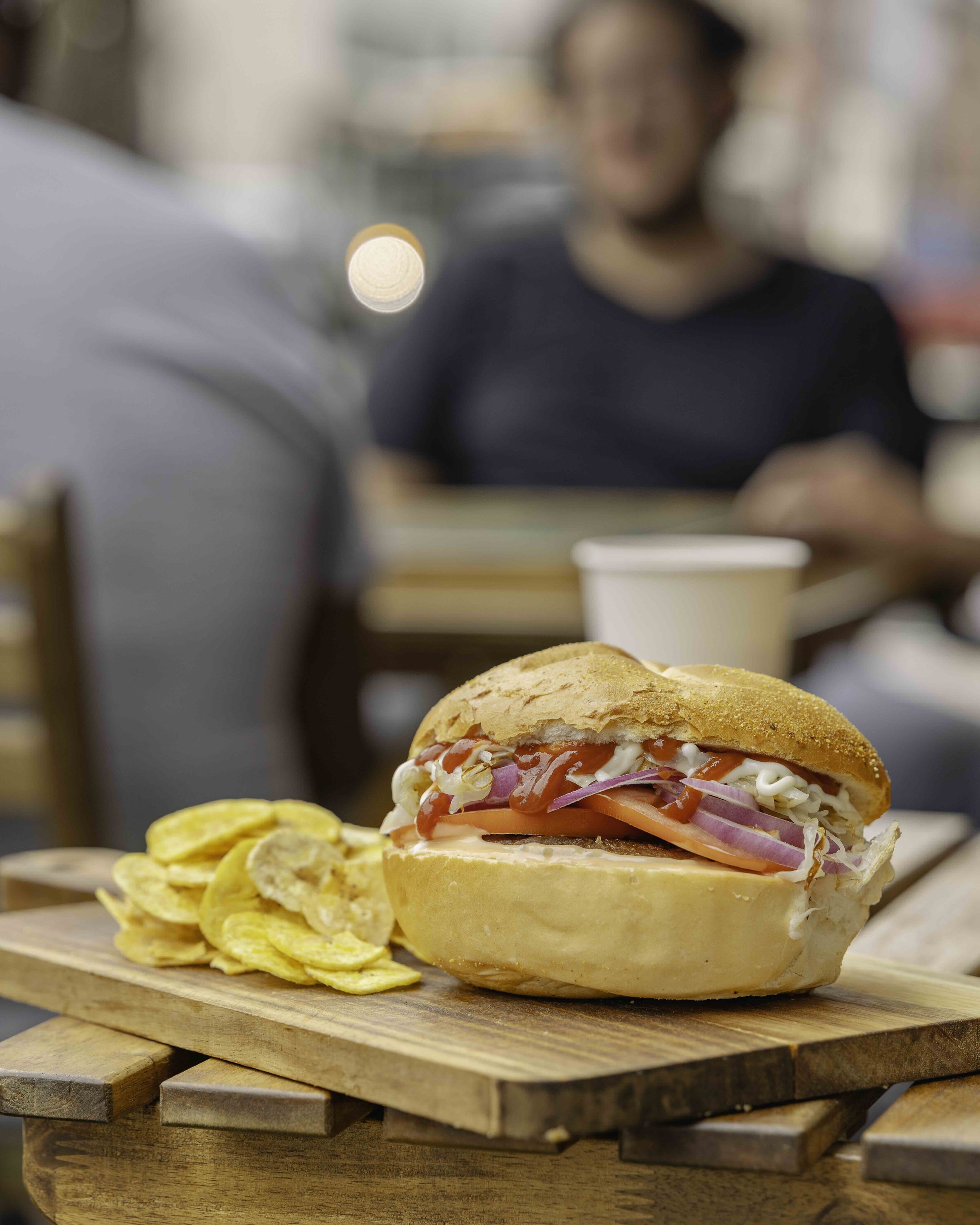 Sandwich with onions, tomatoes, and mayonnaise on a bun, served with potato chips on a wooden tray, with a blurry background of people dining indoors.