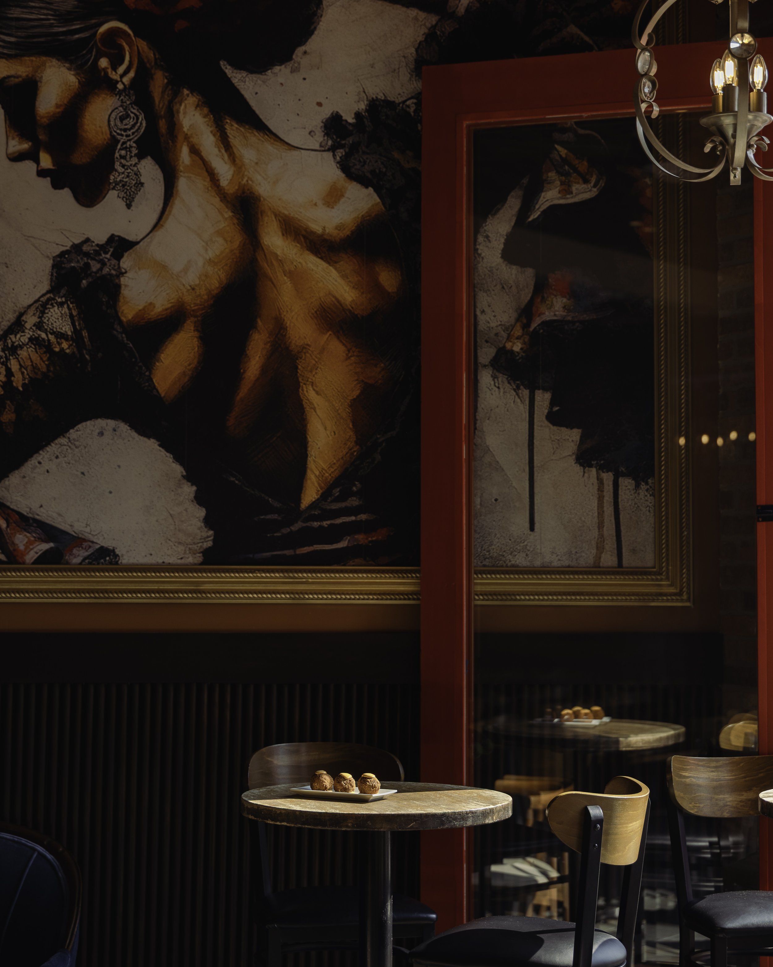 Interior of a restaurant or cafe with round tables and wooden chairs, dessert on a plate on one table, large wall art of a woman's profile, a chandelier, and a red door or partition with glass.