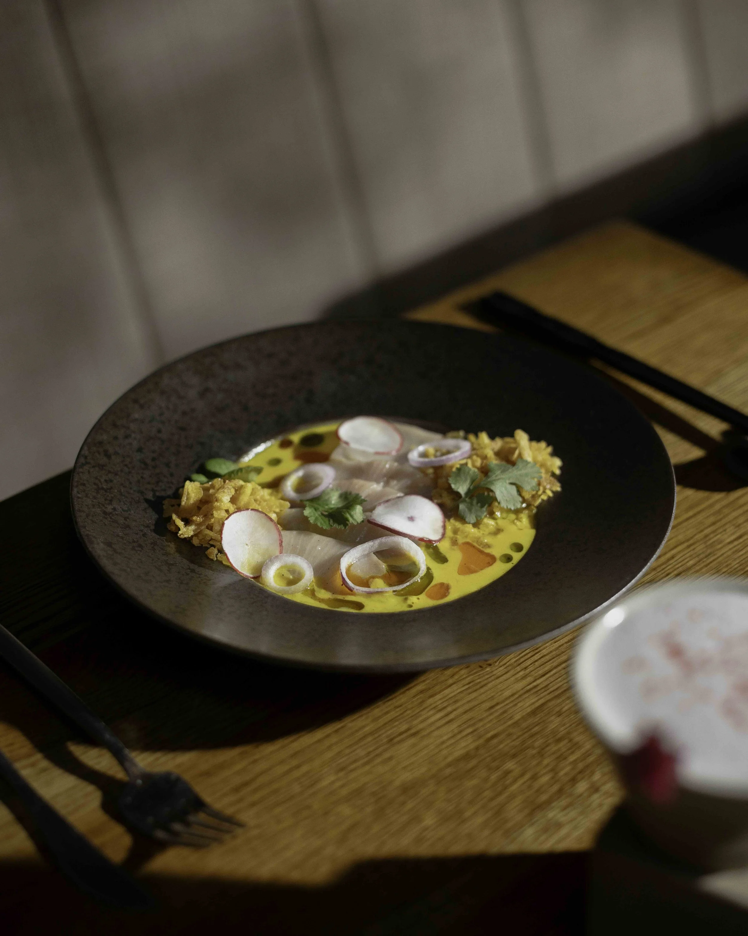 A black plate with yellow curry, rice, sliced radish, sliced onion, cilantro, and chili oil, on a wooden table.