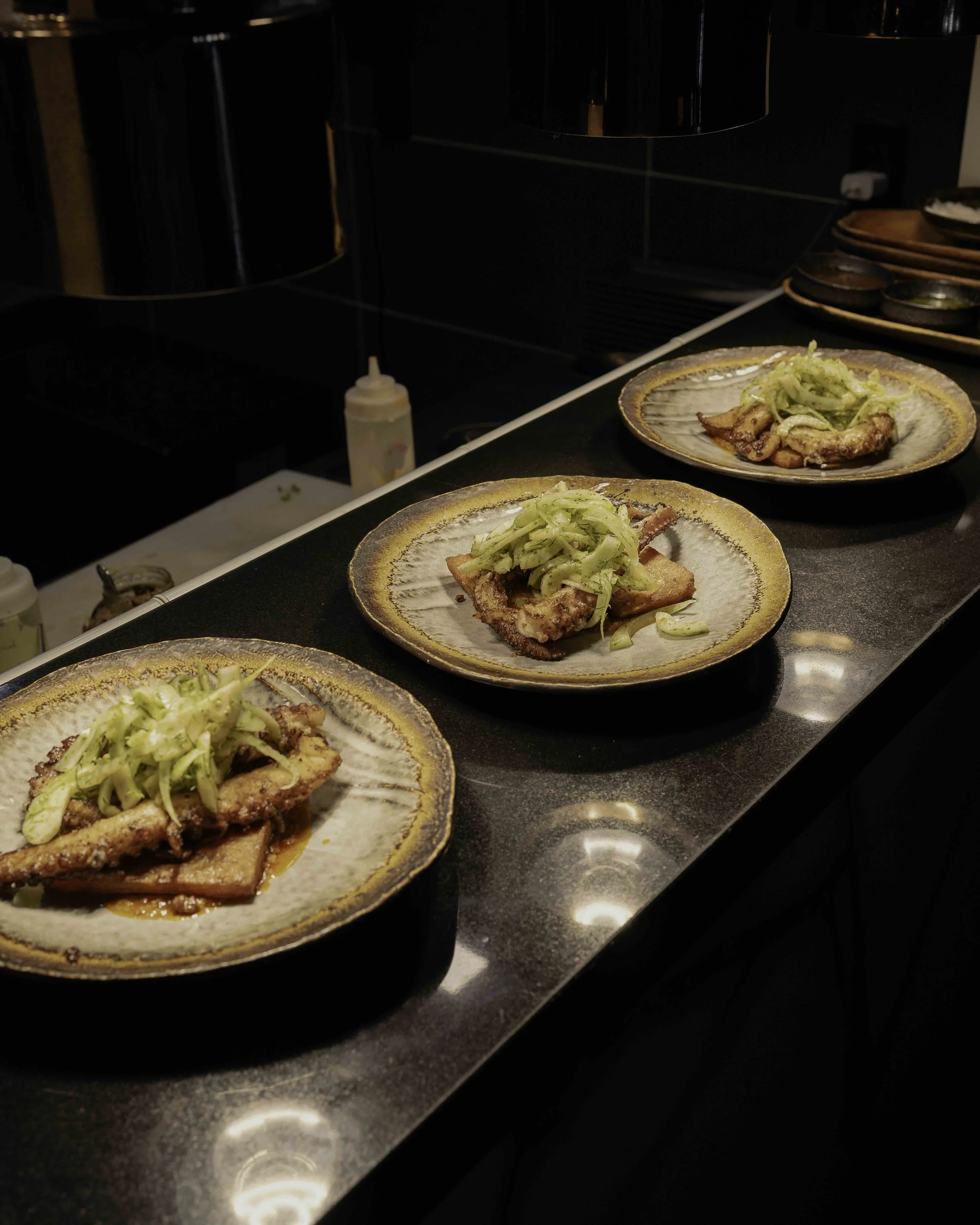 Four plates with grilled fish topped with shredded vegetables on a black countertop in a kitchen.