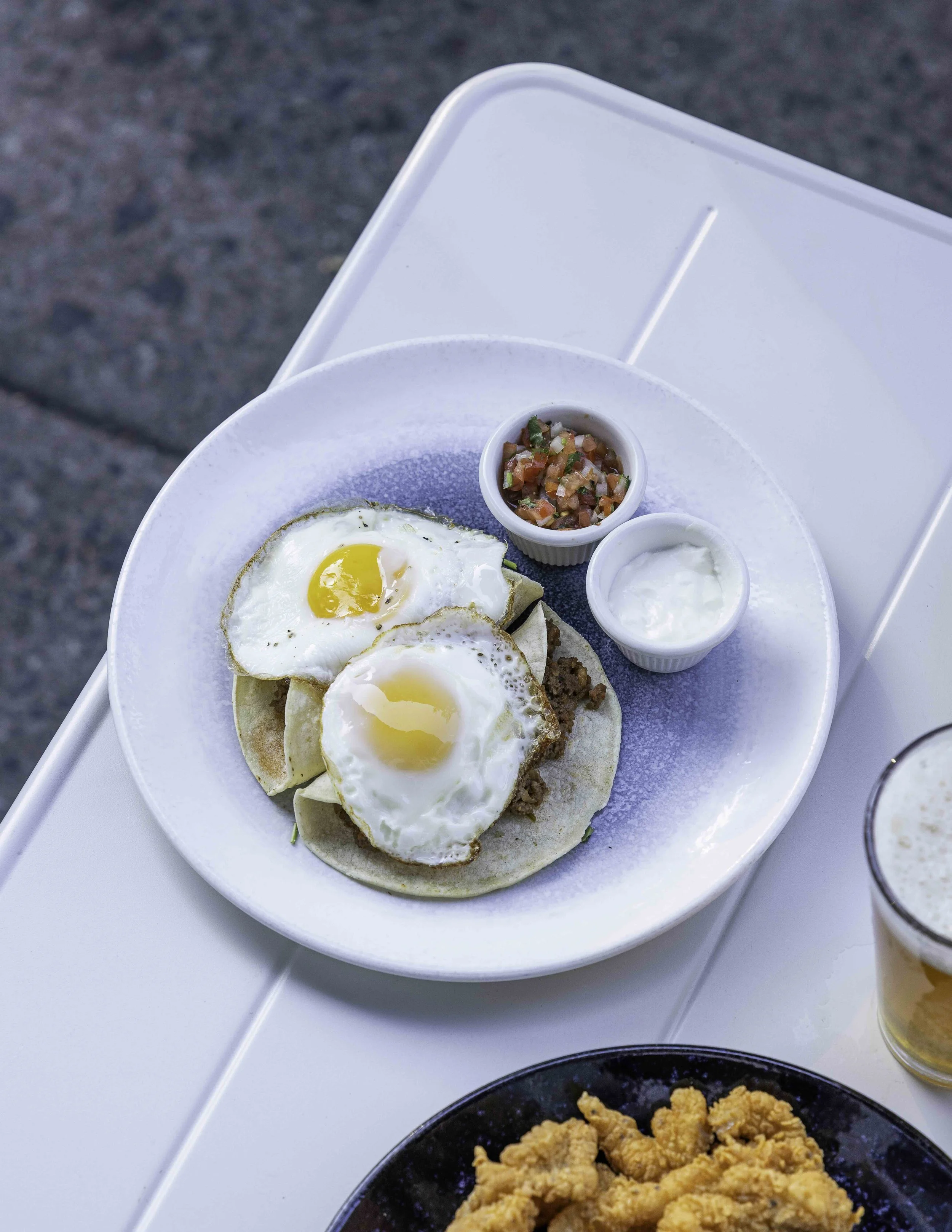 Plate of breakfast tacos topped with fried eggs, accompanied by salsa, sour cream, and a side of fried potatoes, with a glass of beer on the side.