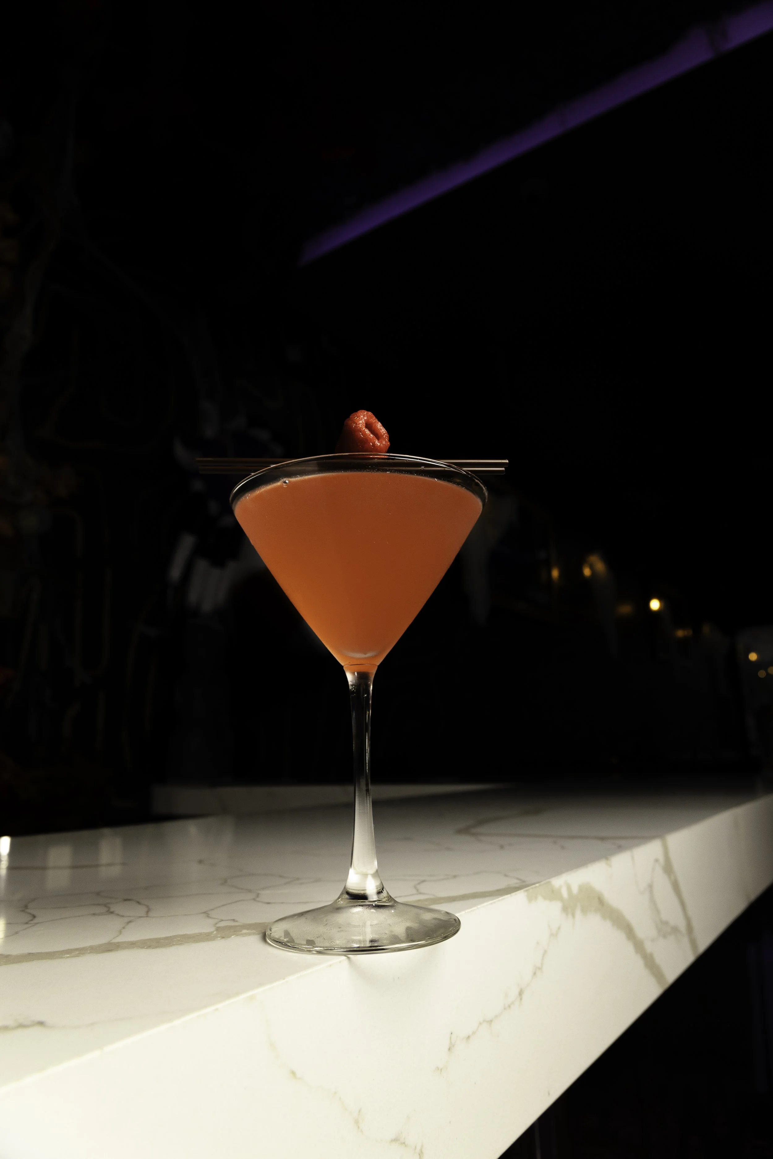 A pink cocktail with a raspberry garnish on top, served in a martini glass on a white marble bar counter in a dimly lit setting.