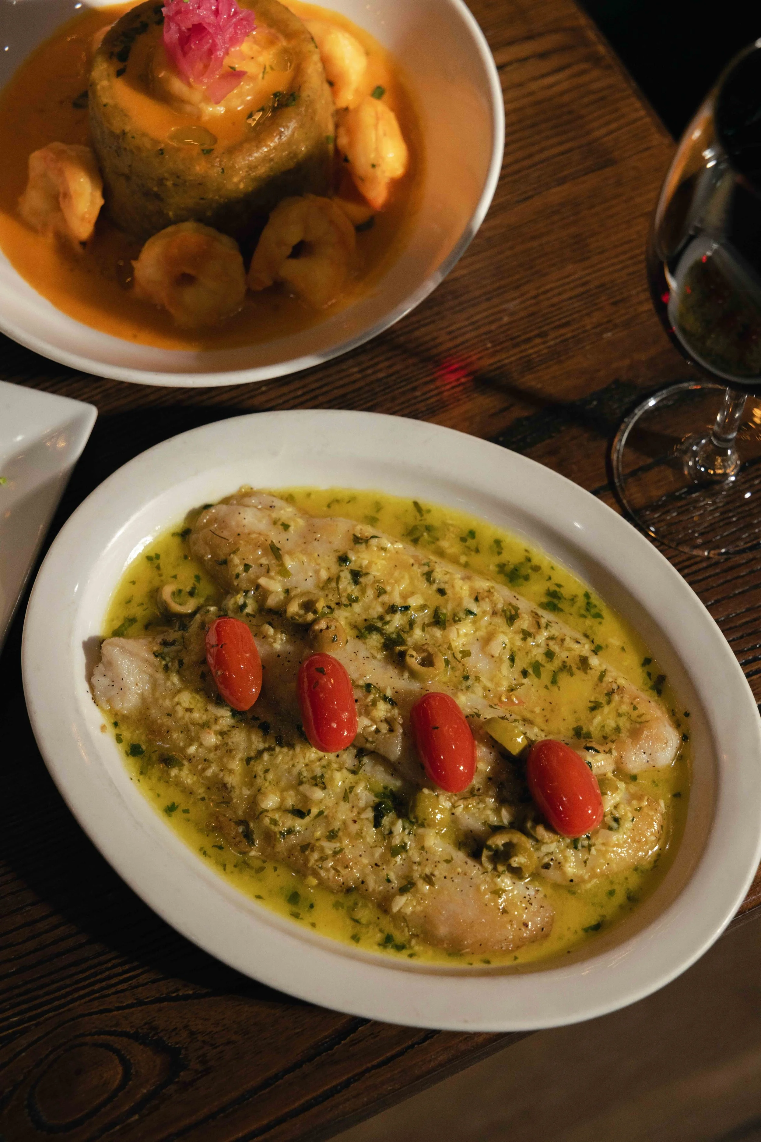 A white oval dish with cooked fish in a green herb sauce garnished with cherry tomatoes. Behind, a bowl with a stuffed potato topped with pink pickled onions and surrounded by shrimp, and a glass of red wine.