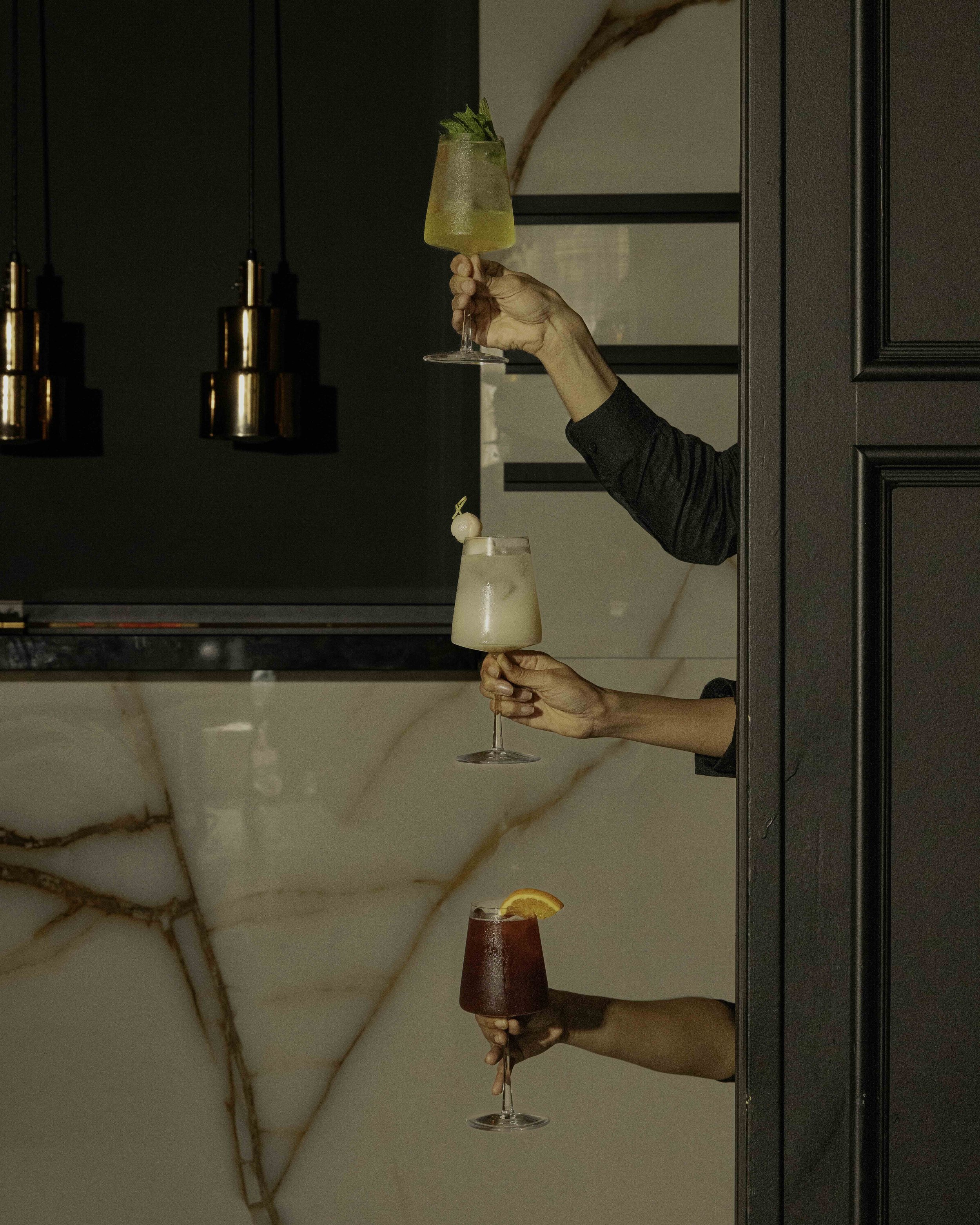 Three people holding cocktails with their arms extended through a wall. The first cocktail is a yellow drink garnished with mint, the second is a creamy white drink with a garnish, and the third is a dark red drink with an orange slice.