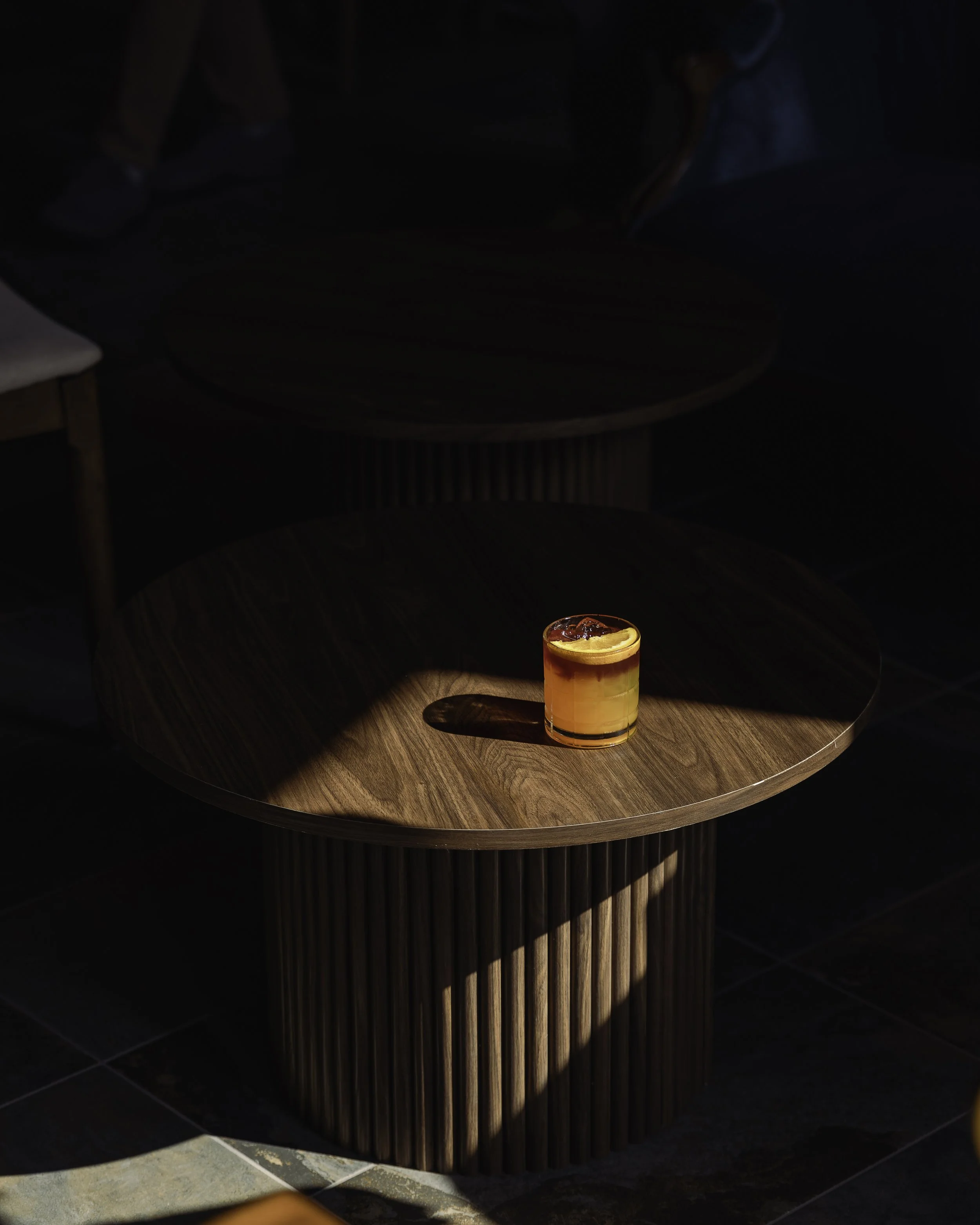 A wooden table with a cocktail glass containing an orange drink topped with fruit, illuminated by a beam of sunlight.