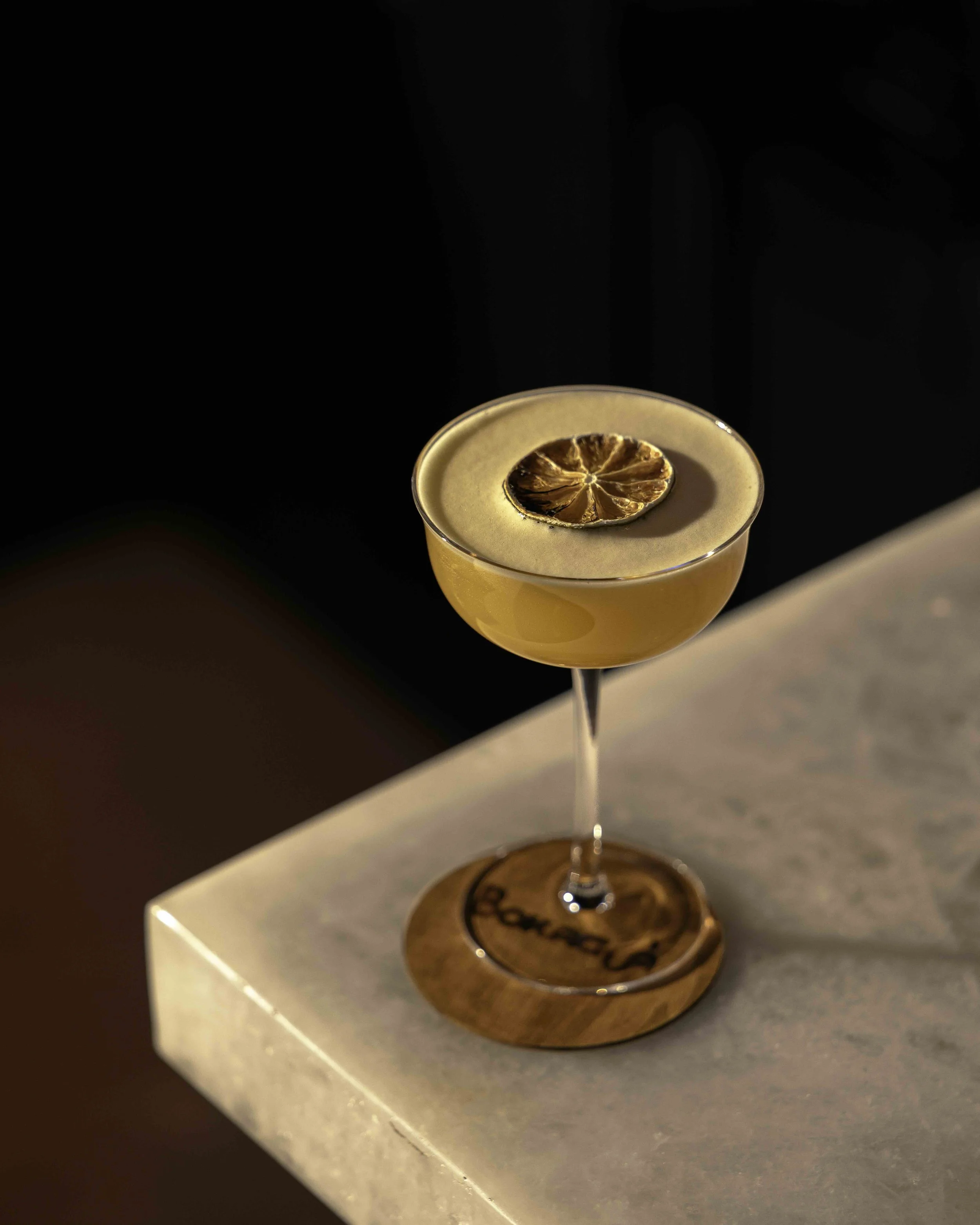 Cocktail in a coupe glass garnished with a dried citrus slice, sitting on a wooden coaster on a marble surface against a dark background.