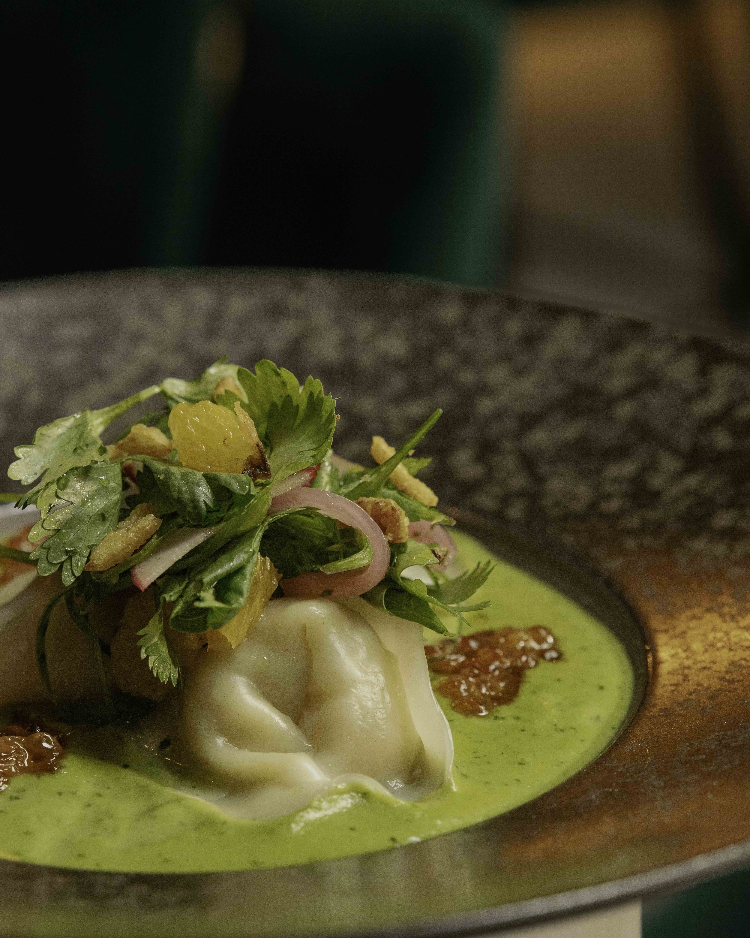 Close-up of a plated dish with dumplings topped with fresh herbs, sliced onions, and yellow fruit, served with a creamy green sauce.