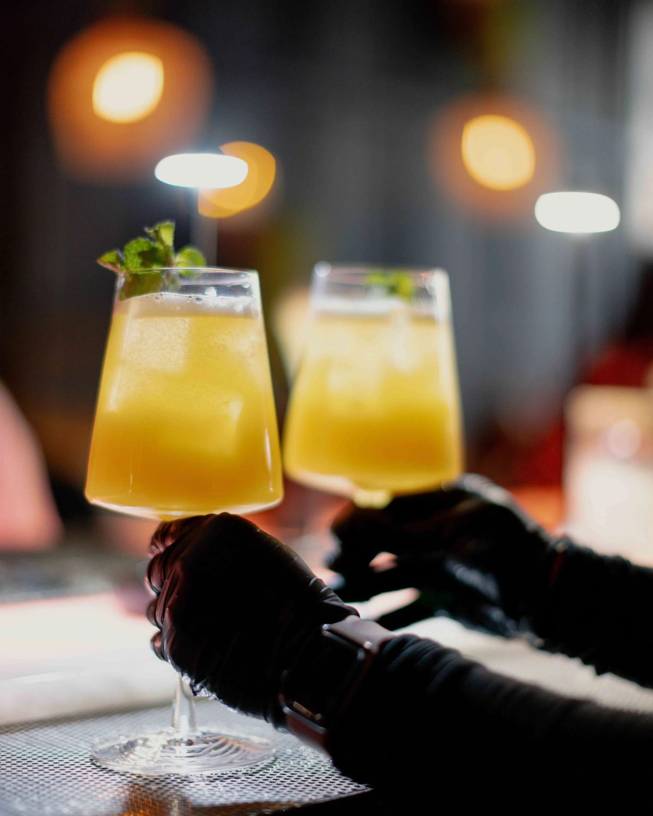 Two hands wearing black gloves are holding glasses of yellow cocktails garnished with mint leaves in a bar or restaurant setting with bokeh lights in the background.