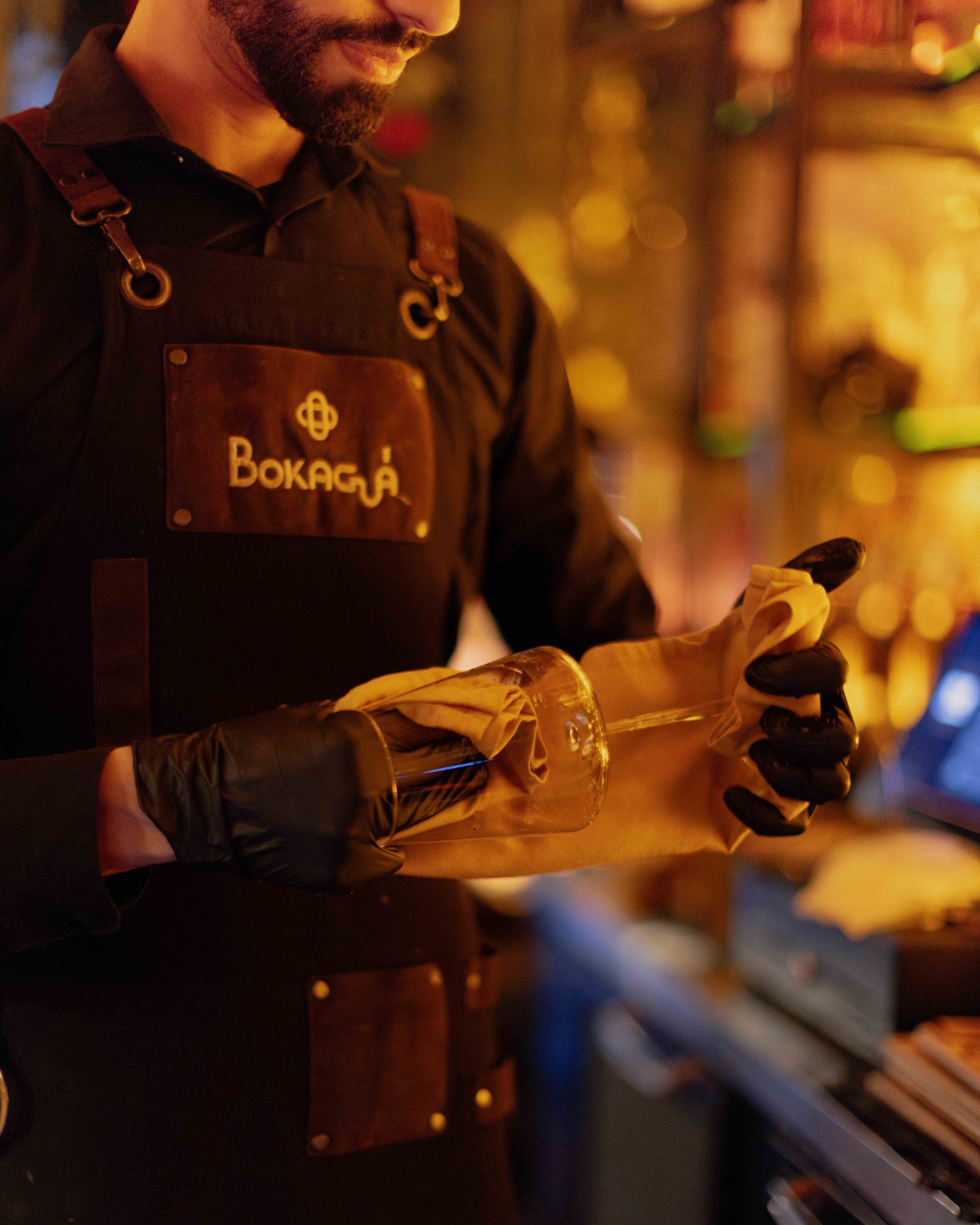 A bartender wearing a black apron with the word 'Bokajá' and gloves is holding a glass bottle wrapped in a cloth, preparing a drink in a dimly lit bar with warm lighting.