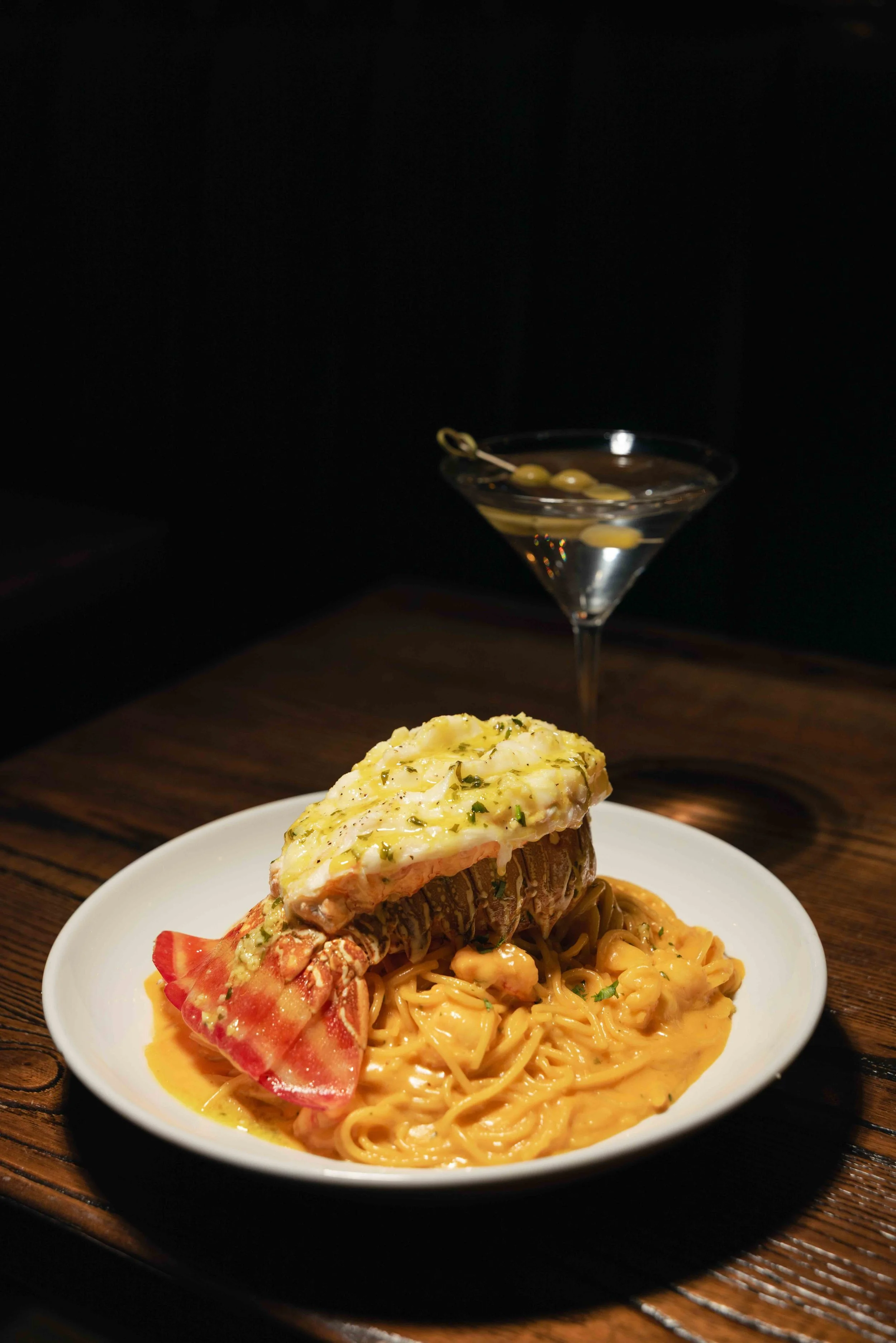 Lobster served over pasta with creamy sauce on a white plate, accompanied by a martini with olives in a glass, on a dark wooden table.