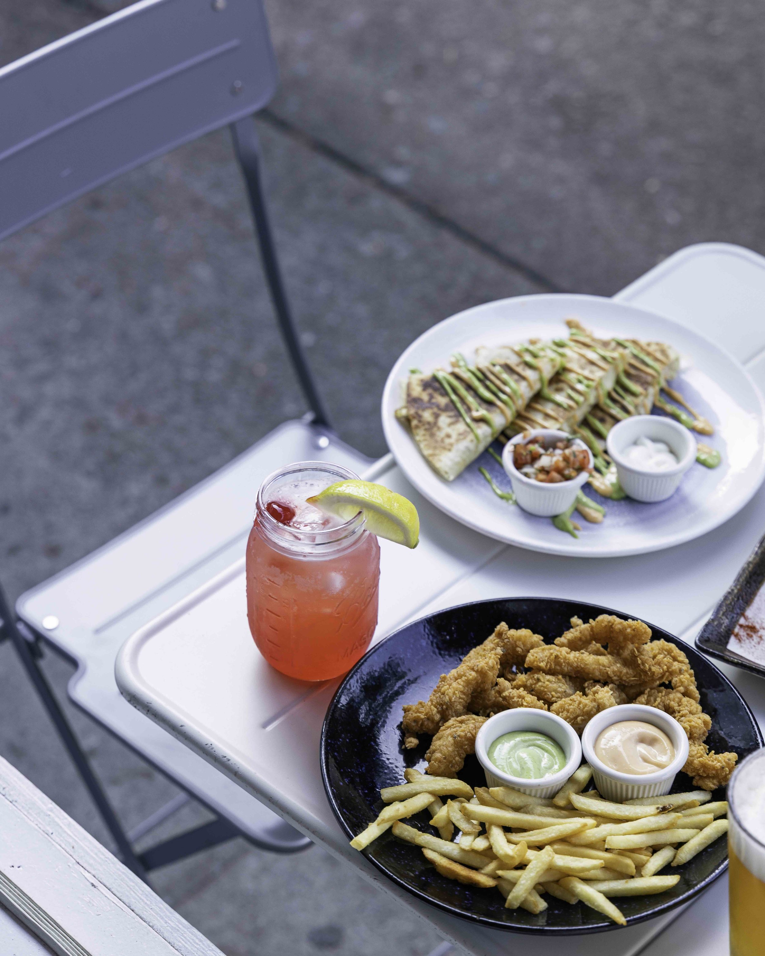 A table with a plate of fried chicken tenders, French fries with two dipping sauces, a drink with a lemon wedge, and a plate of quesadilla with sides of salsa, sour cream, and guacamole.