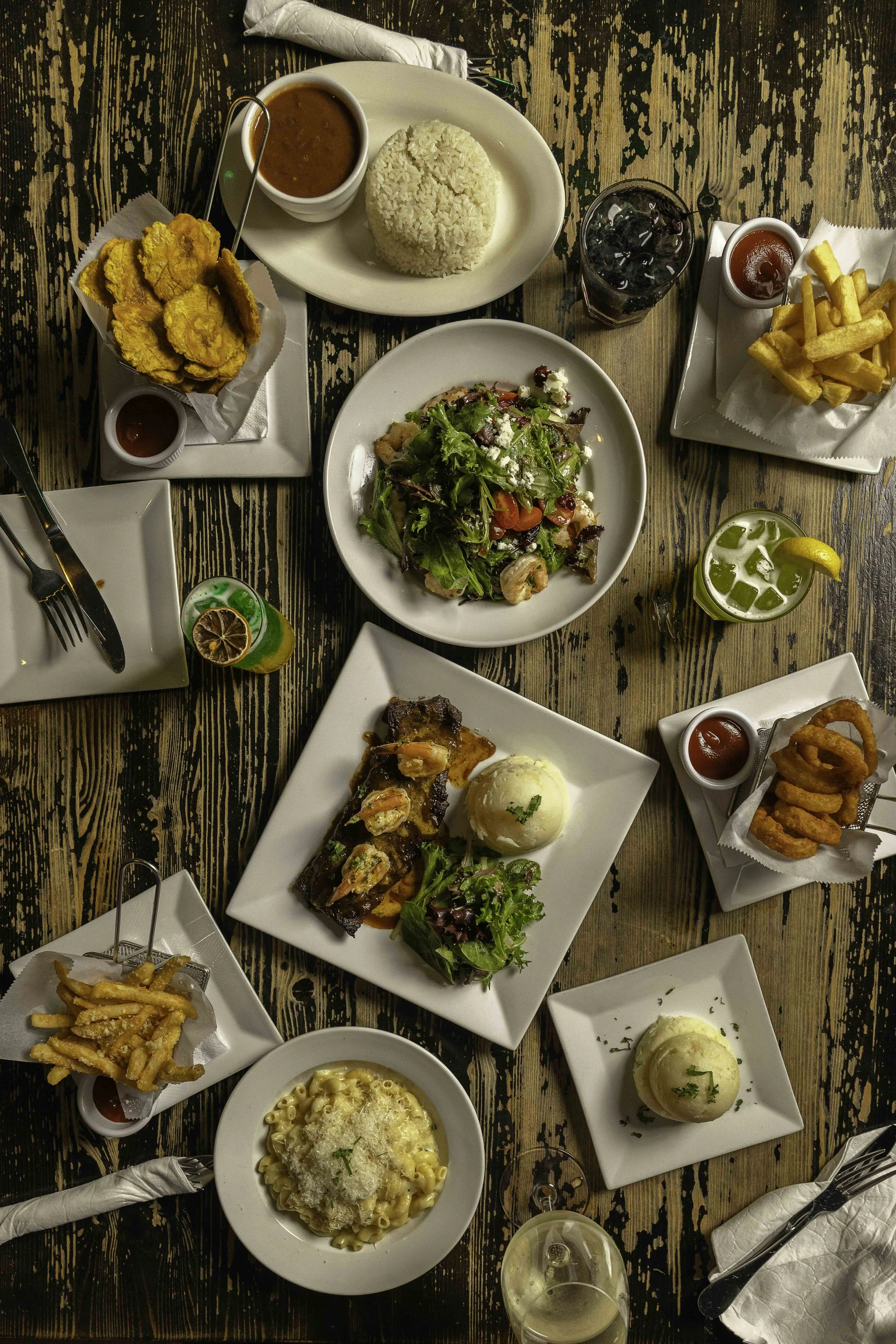 Assorted dishes on a rustic wooden table including salad, fried plantains, french fries, onion rings, macaroni and cheese, grilled meat with shrimp, rice, and various drinks.