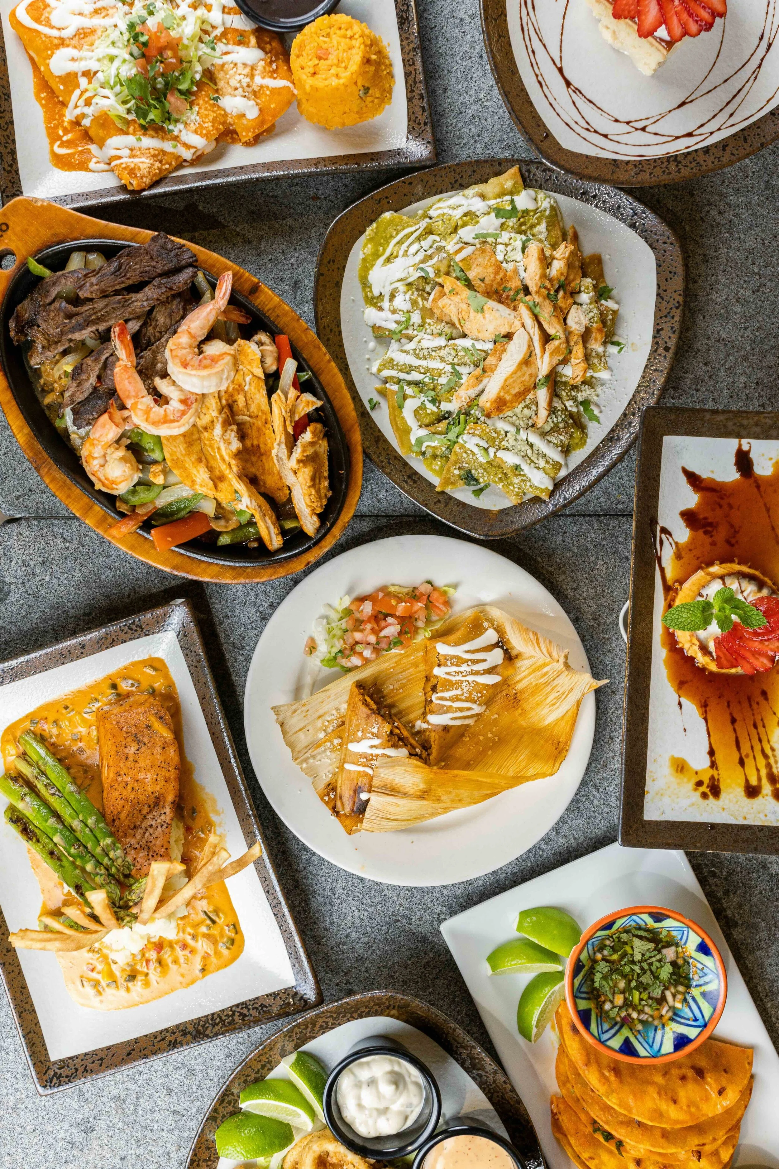 A variety of Mexican dishes including enchiladas, tacos, grilled meat with vegetables, a quesadilla, and desserts such as flan and strawberry-topped cheesecake, served on plates and bowls on a gray table.