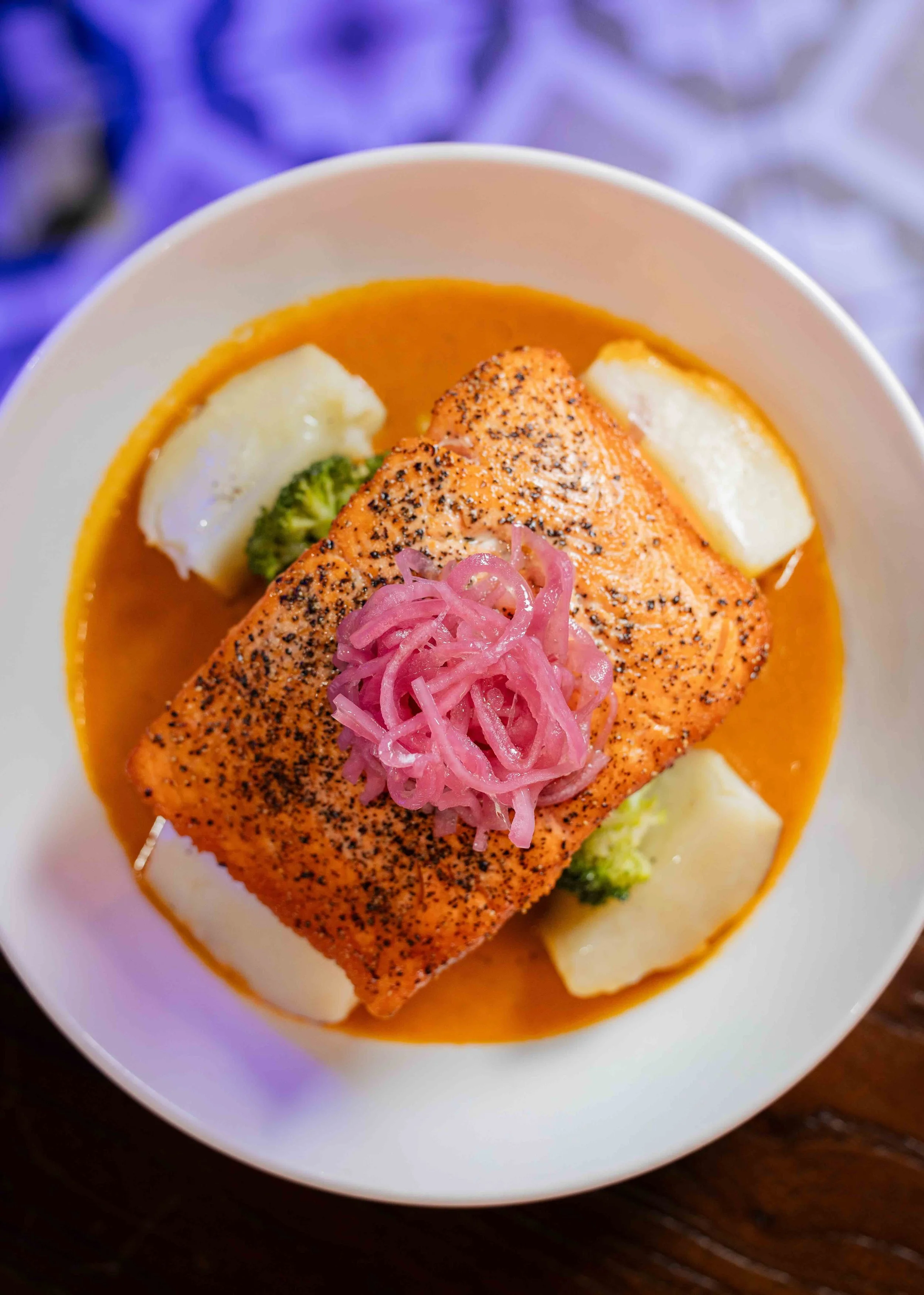 A bowl of cooked salmon topped with pickled onions, served over a bed of mashed potatoes and green vegetables in an orange sauce.