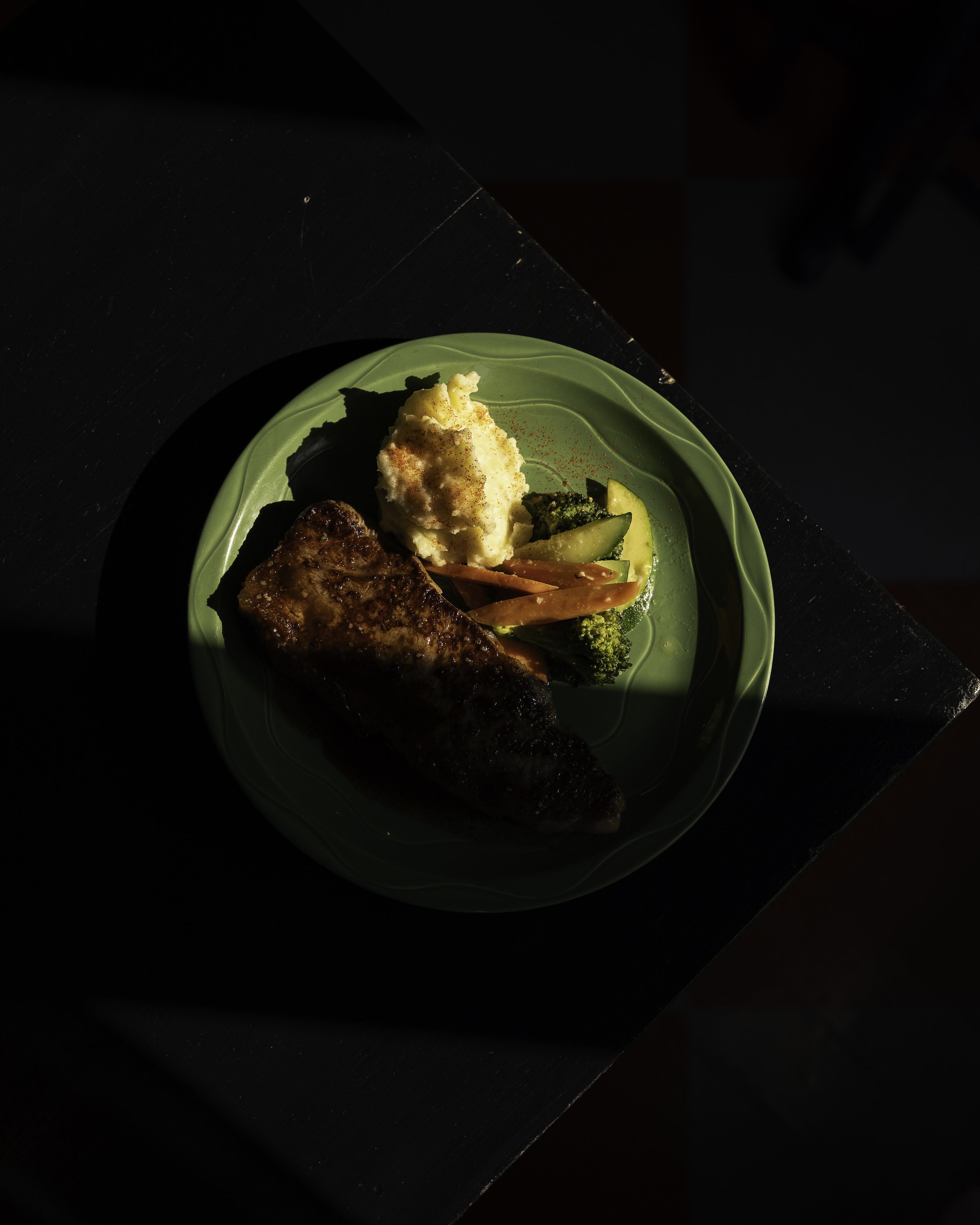 Plate of grilled meat, mashed potatoes, and mixed vegetables on a green plate with a dark background.