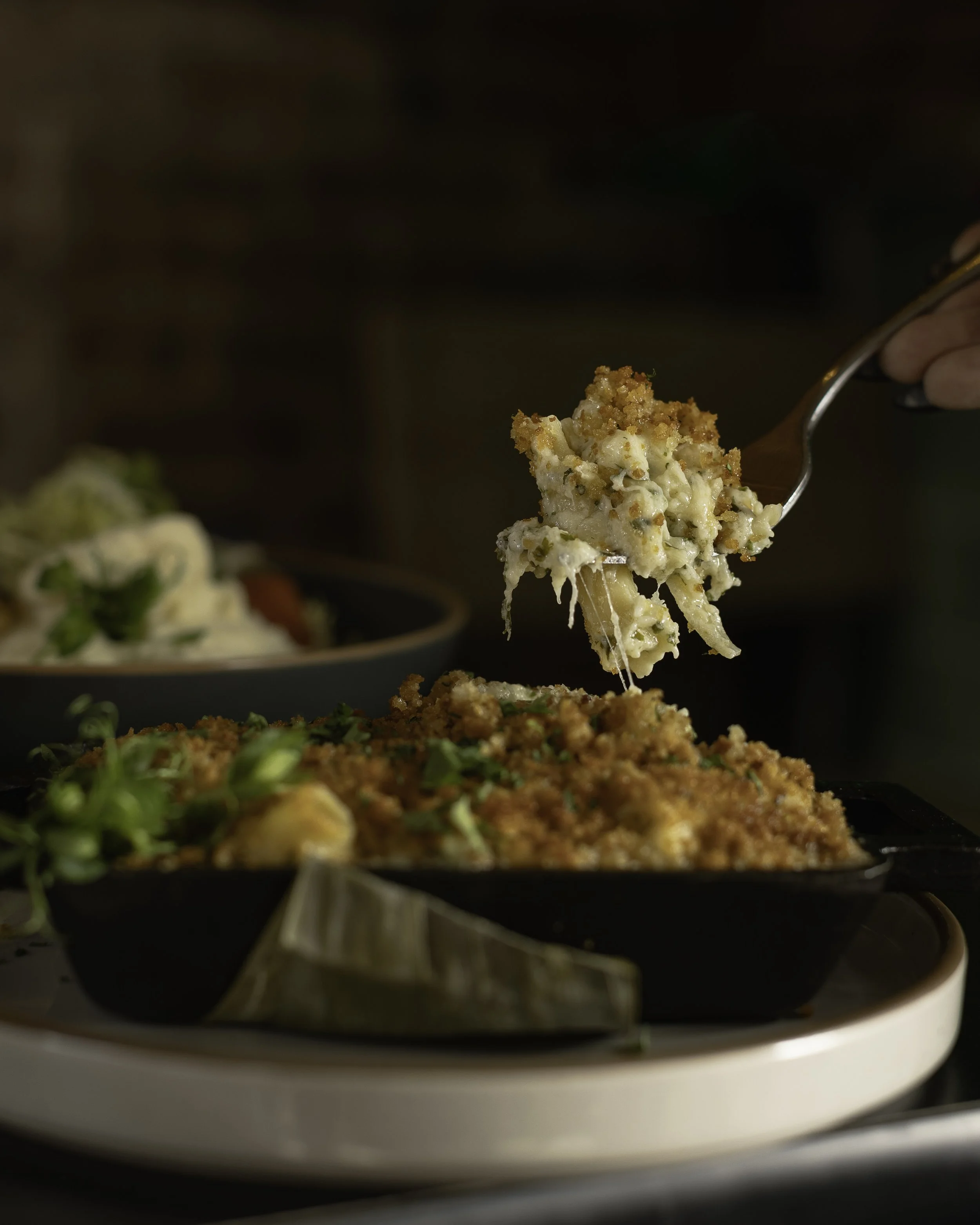 A serving spoon lifting a portion of baked pasta casserole with cheese and breadcrumb topping, with another dish of pasta salad in the background.
