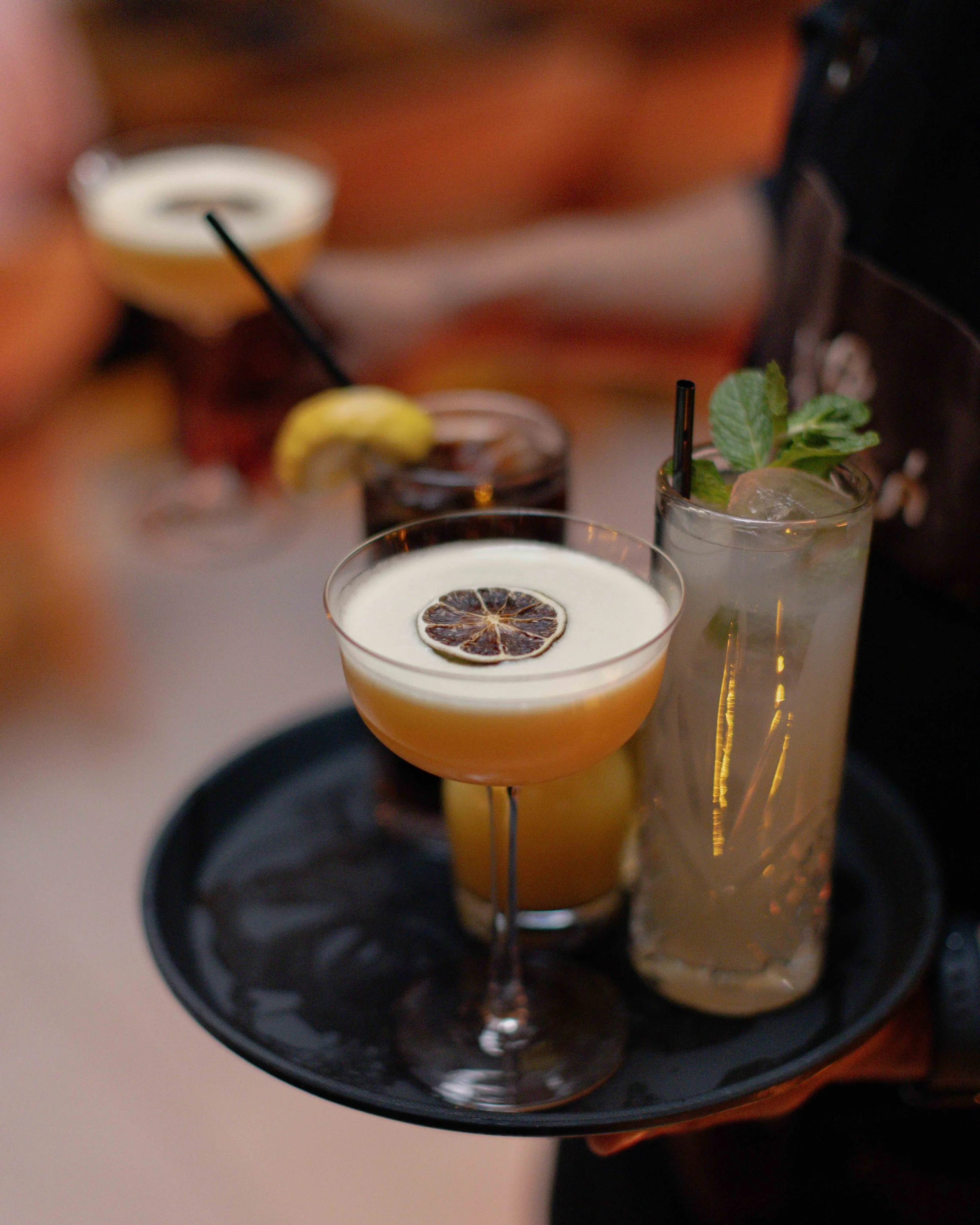 A black tray holding four cocktail glasses with garnishes, featuring dried citrus slices and mint leaves.