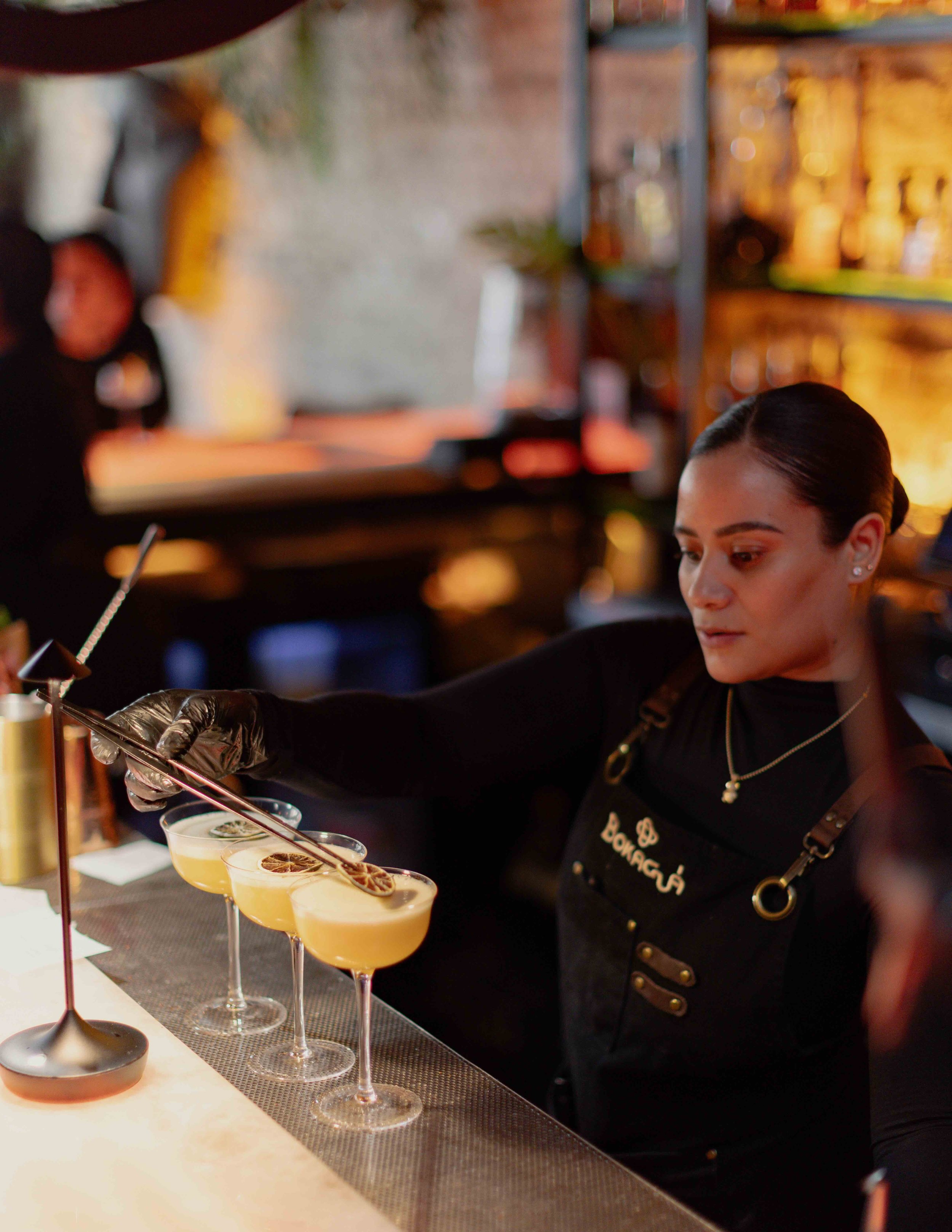 A bartender in black attire and gloves preparing cocktails at a bar, with multiple cocktails garnished with lemon slices on the bar counter.