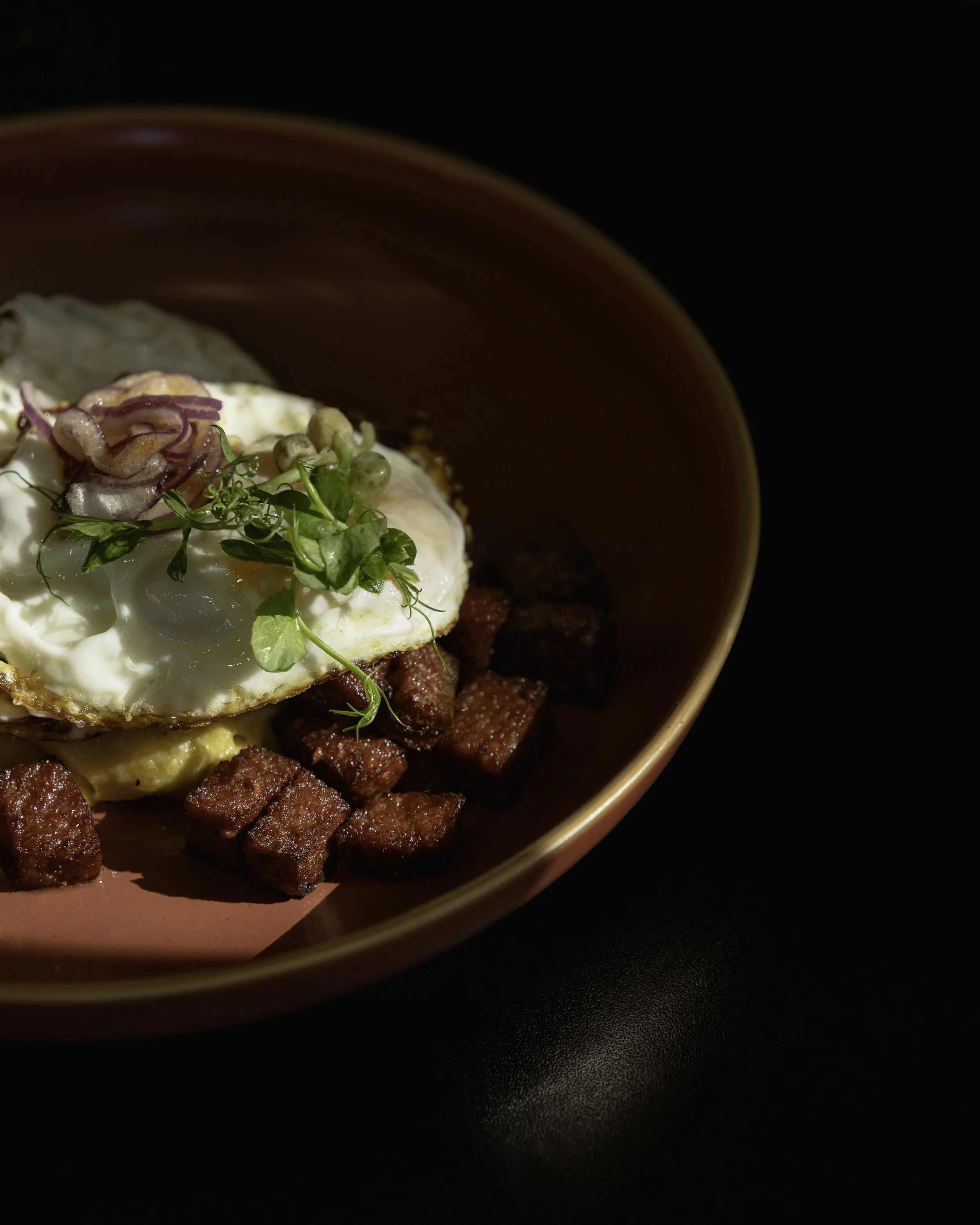 A bowl with diced cooked meat, a fried egg with a runny yolk, garnished with microgreens, served with caramelized onions on top, in a dark setting.