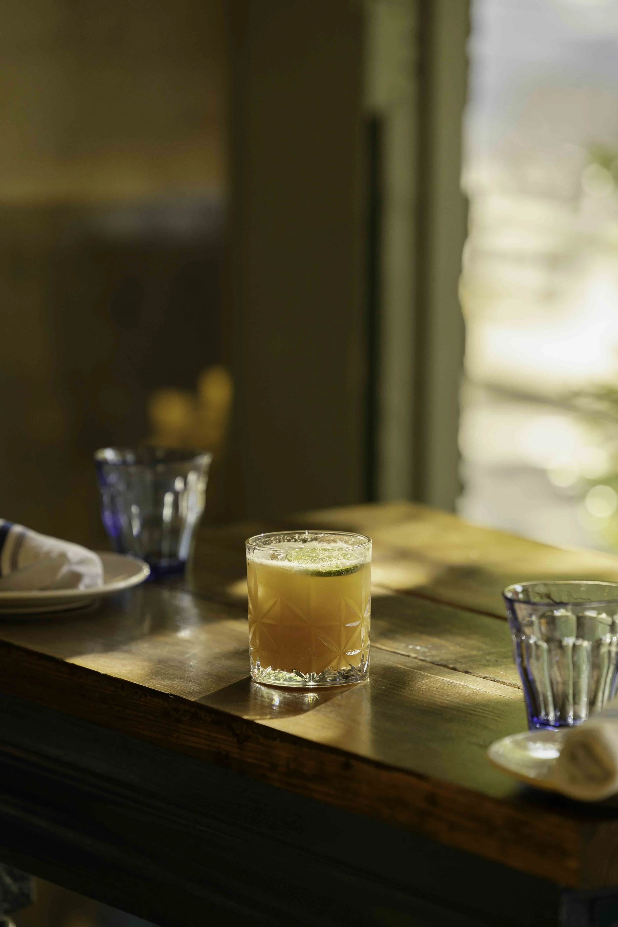 A wooden table with a cocktail garnished with lime, a blue glass, and a white napkin with an app icon in the background.