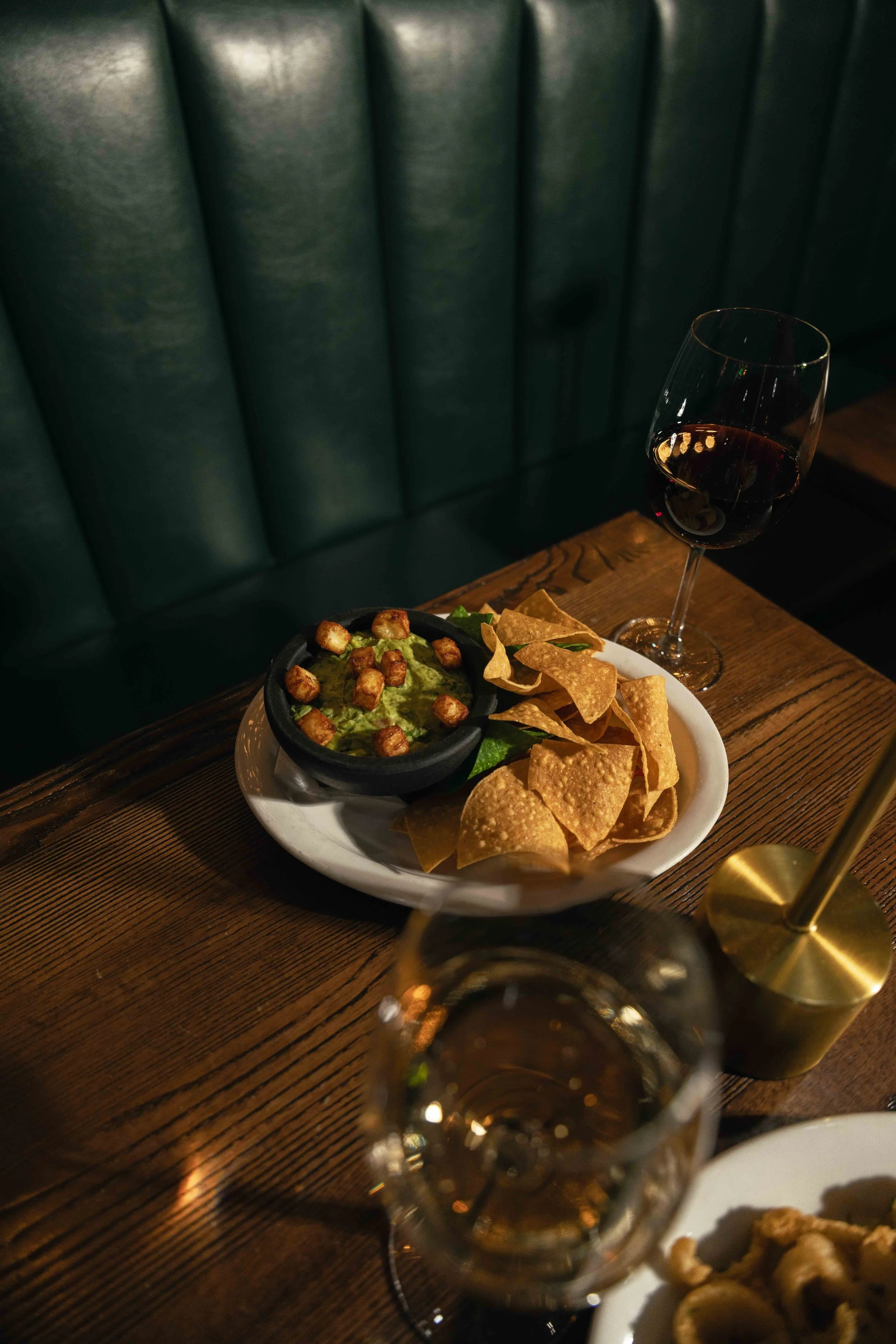 A wooden table with a white plate of tortilla chips, a bowl of guacamole topped with small crispy toppings, and two glasses of red wine in a dimly lit setting.