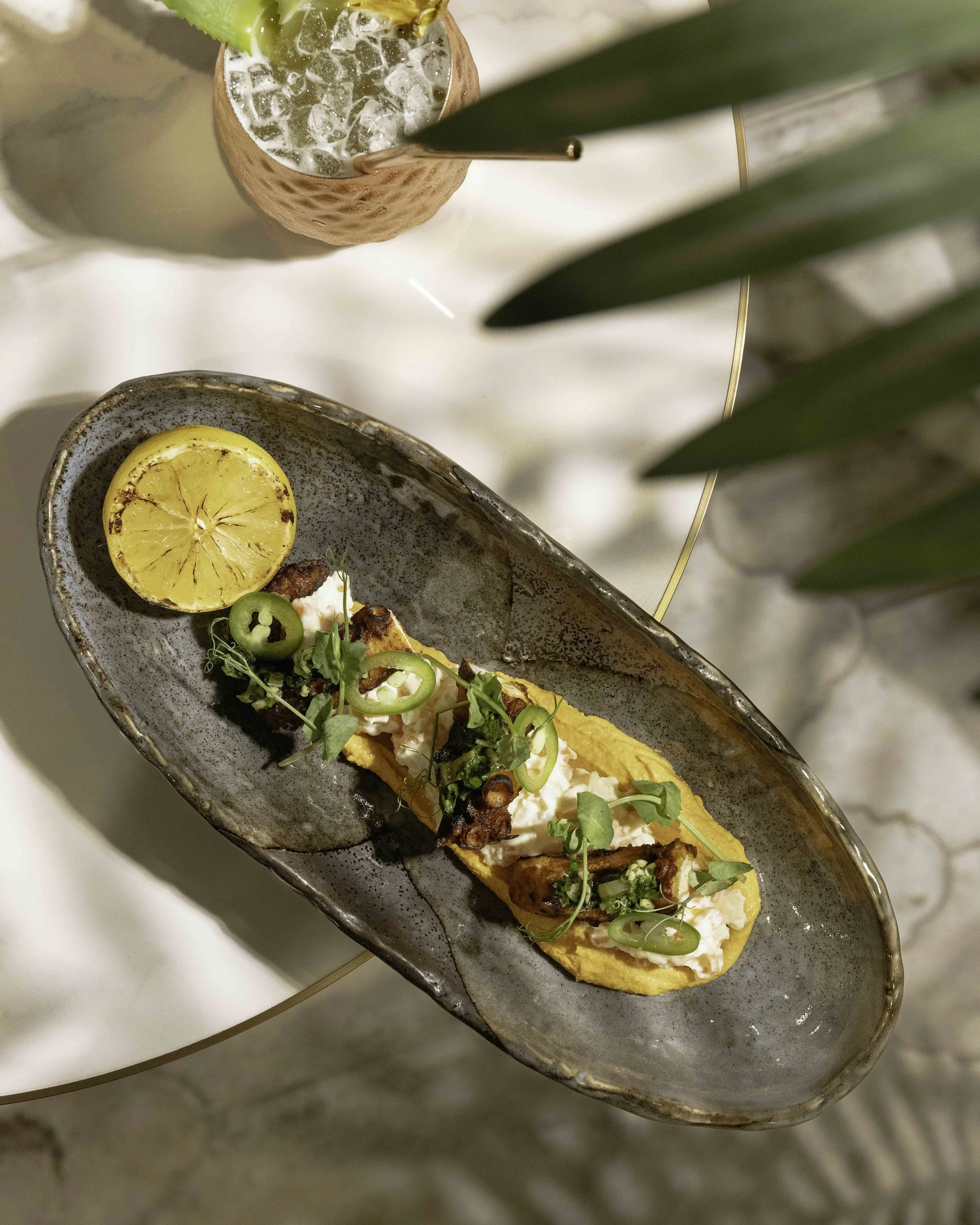 A plate of Mexican street corn topped with crema, cilantro, and jalapeño slices, with a charred lemon half on the side, on a ceramic dish.