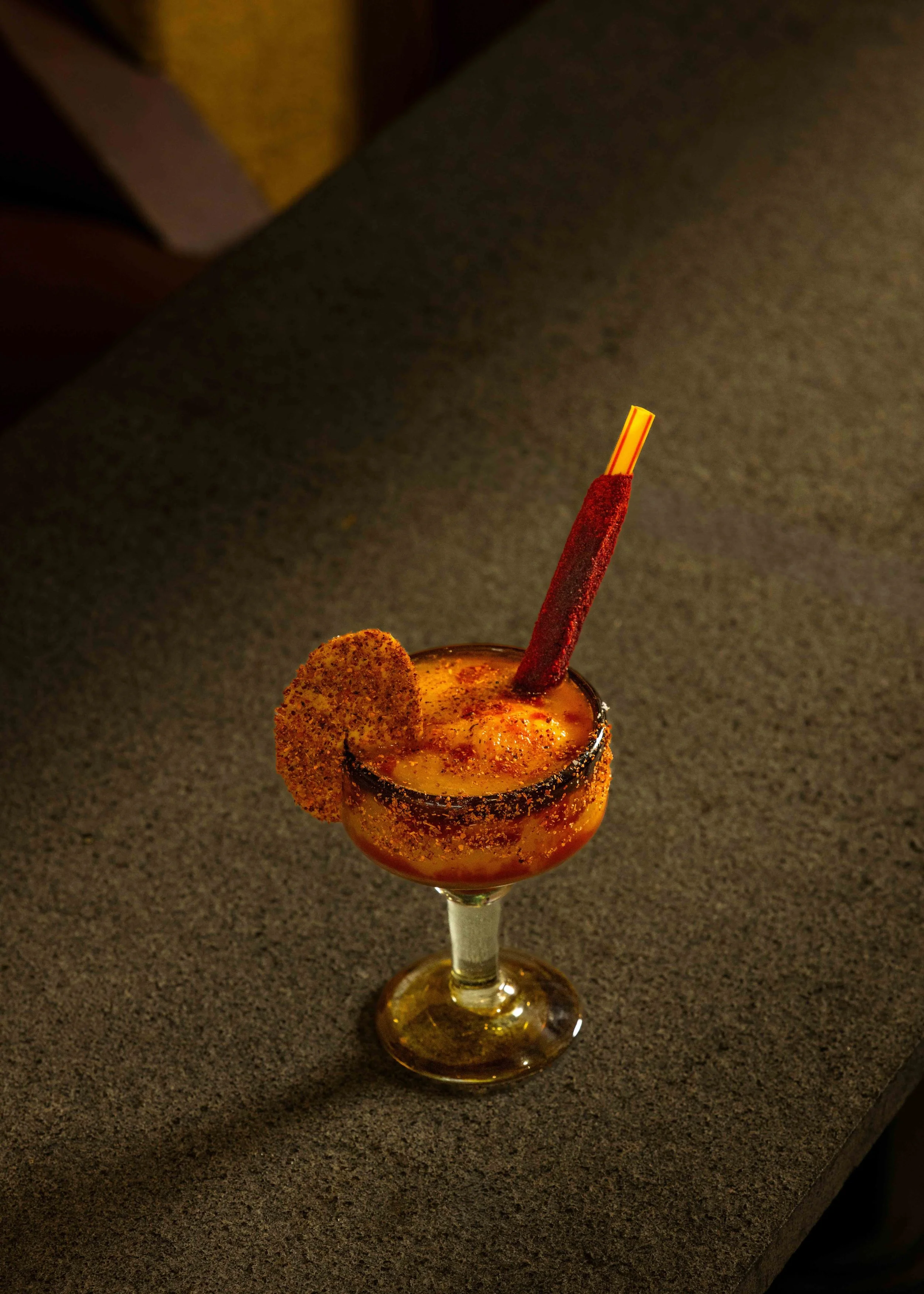 A spicy cocktail in a glass with a salted rim, garnished with a slice of seasoned rim and a red chili pepper straw.