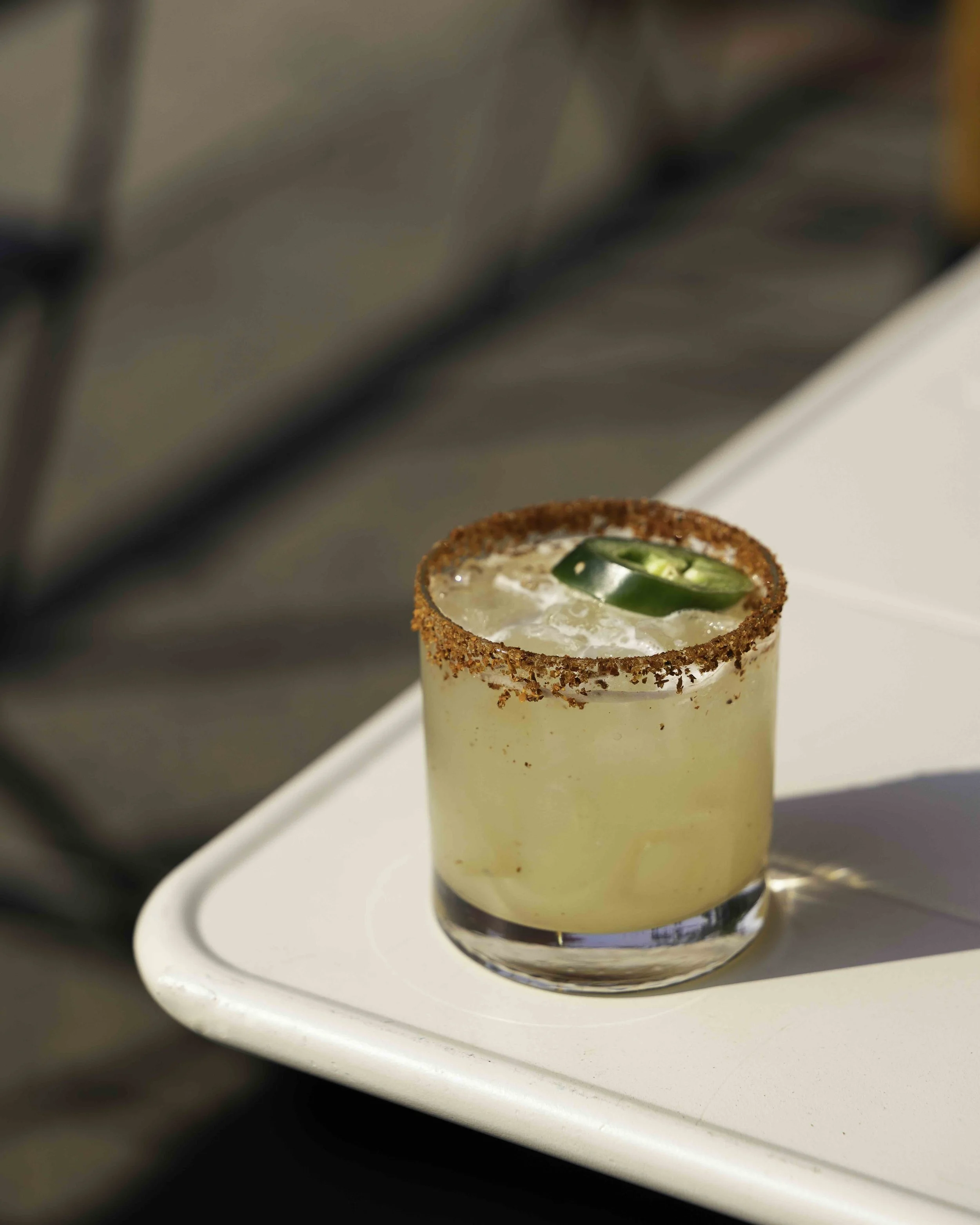 A yellow cocktail in a short glass with a salted rim and sliced jalapeño on top, placed on a white tray.