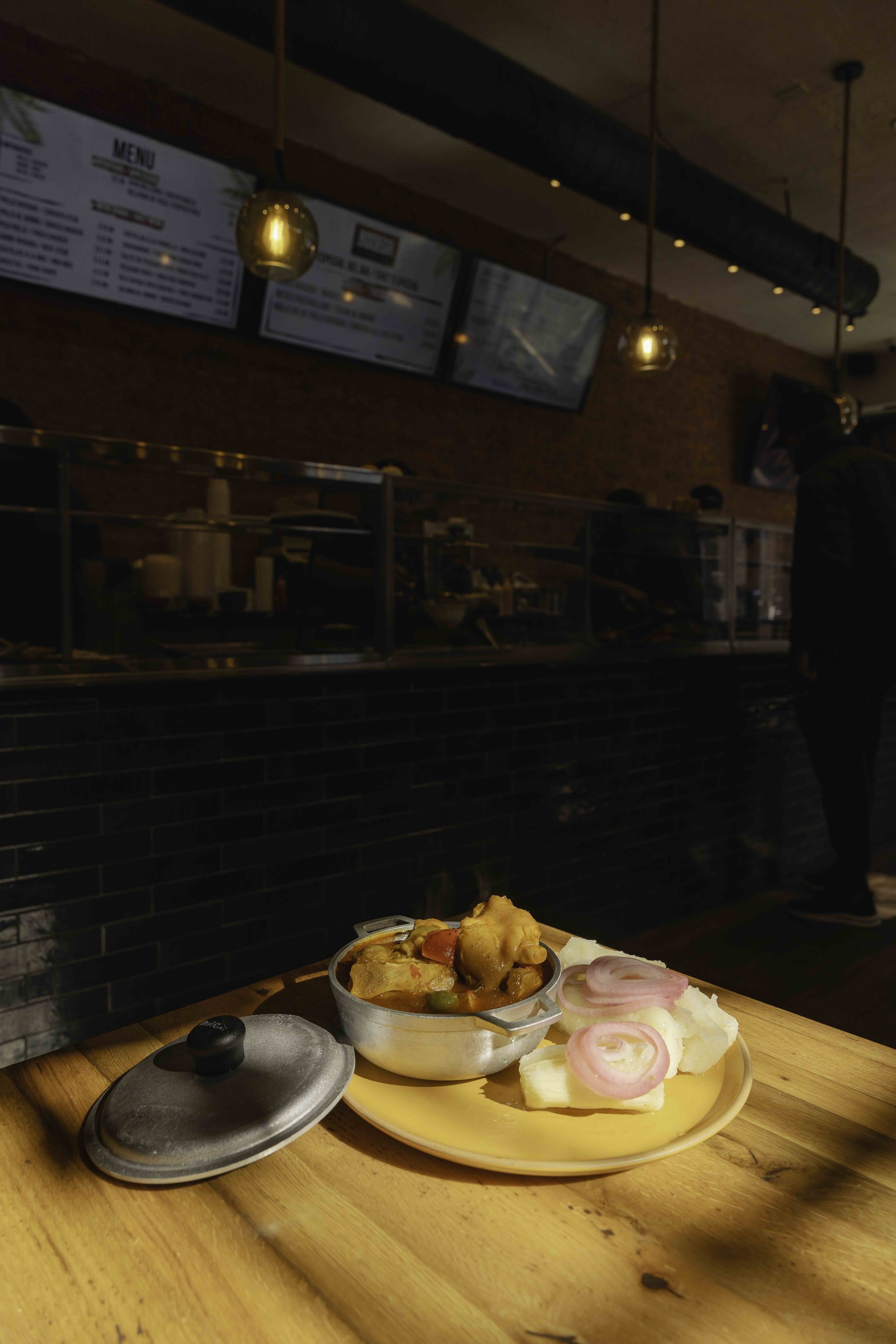 A plate of food with a small pot of stew, pickled onions, and mashed potatoes on a wooden table inside a restaurant.