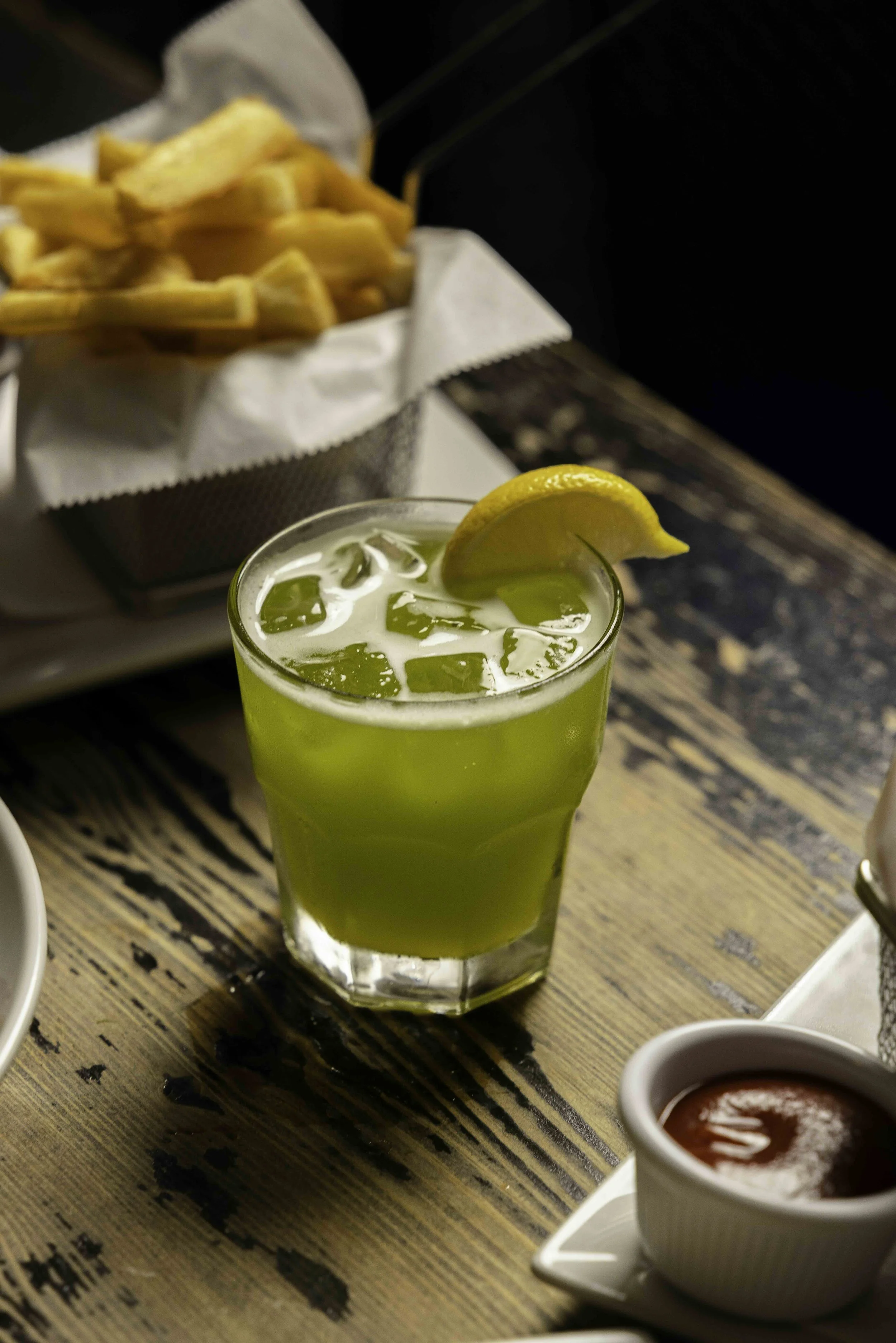 A glass of green iced drink with a lemon wedge garnish on a wooden table, with a basket of French fries in the background and a small bowl of ketchup nearby.