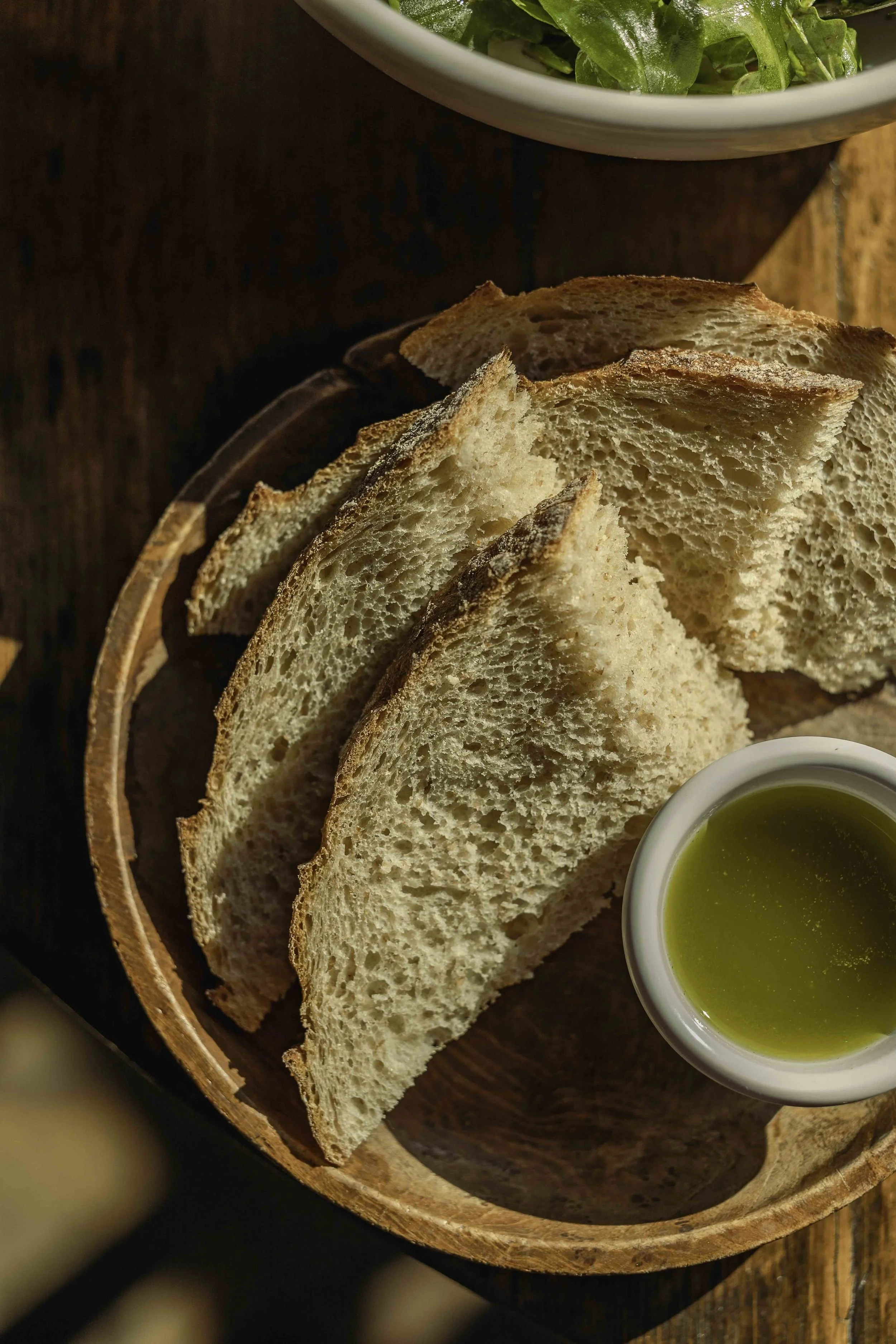 Slices of bread on a wooden plate with a small dish of olive oil, with a bowl of green salad in the background.