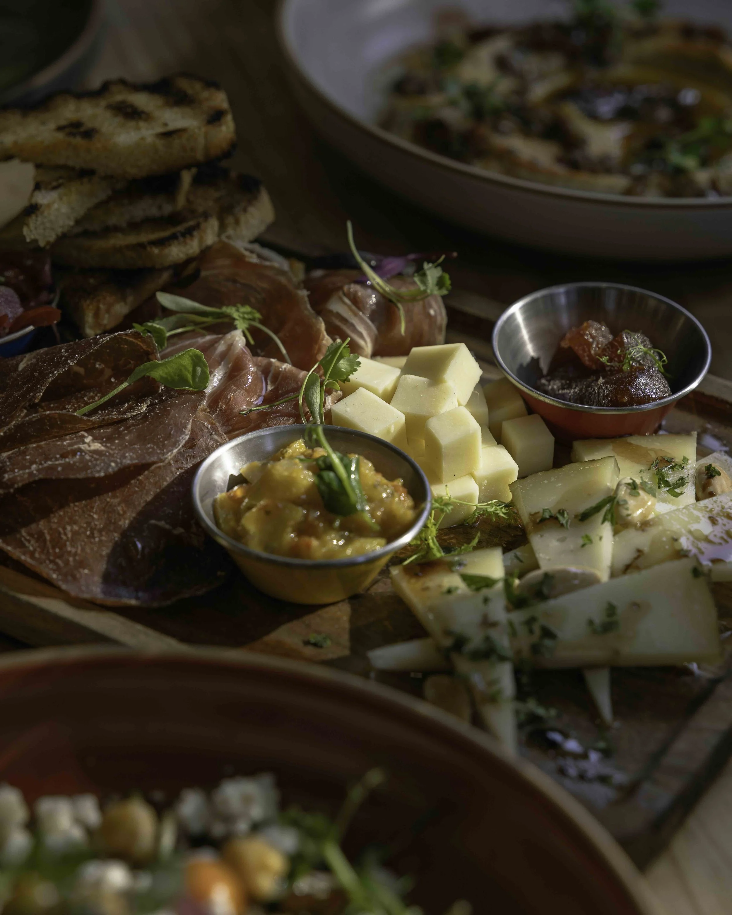 A cheese and charcuterie board with cubed cheese, slices of meat, small bowls of spreads and dips, toasted bread, and garnishes.