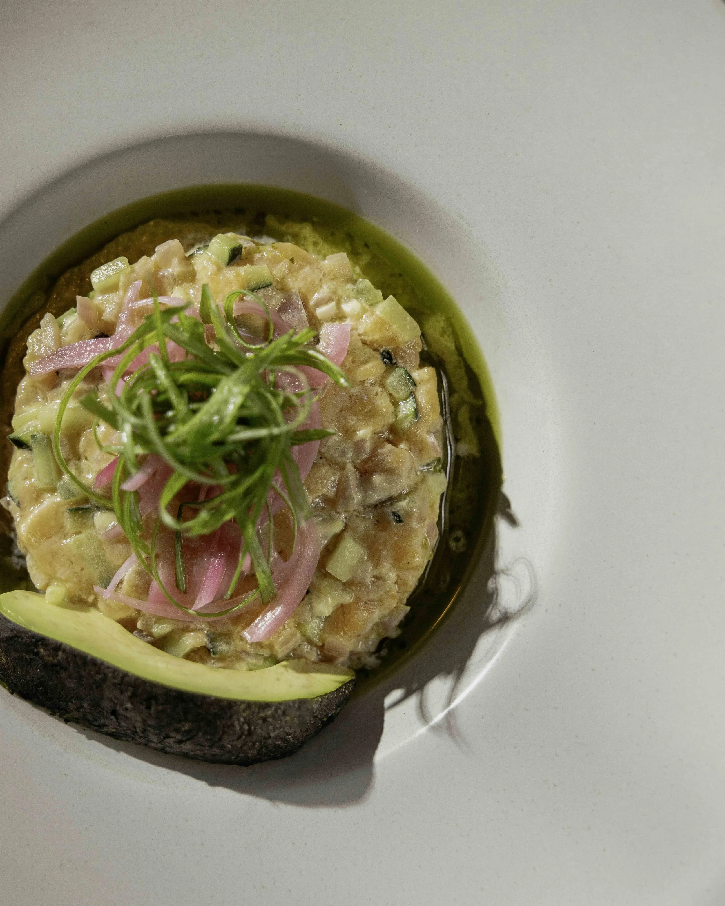 Food dish with a bowl of salmon tartare garnished with green onion curls and pink pickled onion slices, served with a slice of avocado on a white plate.