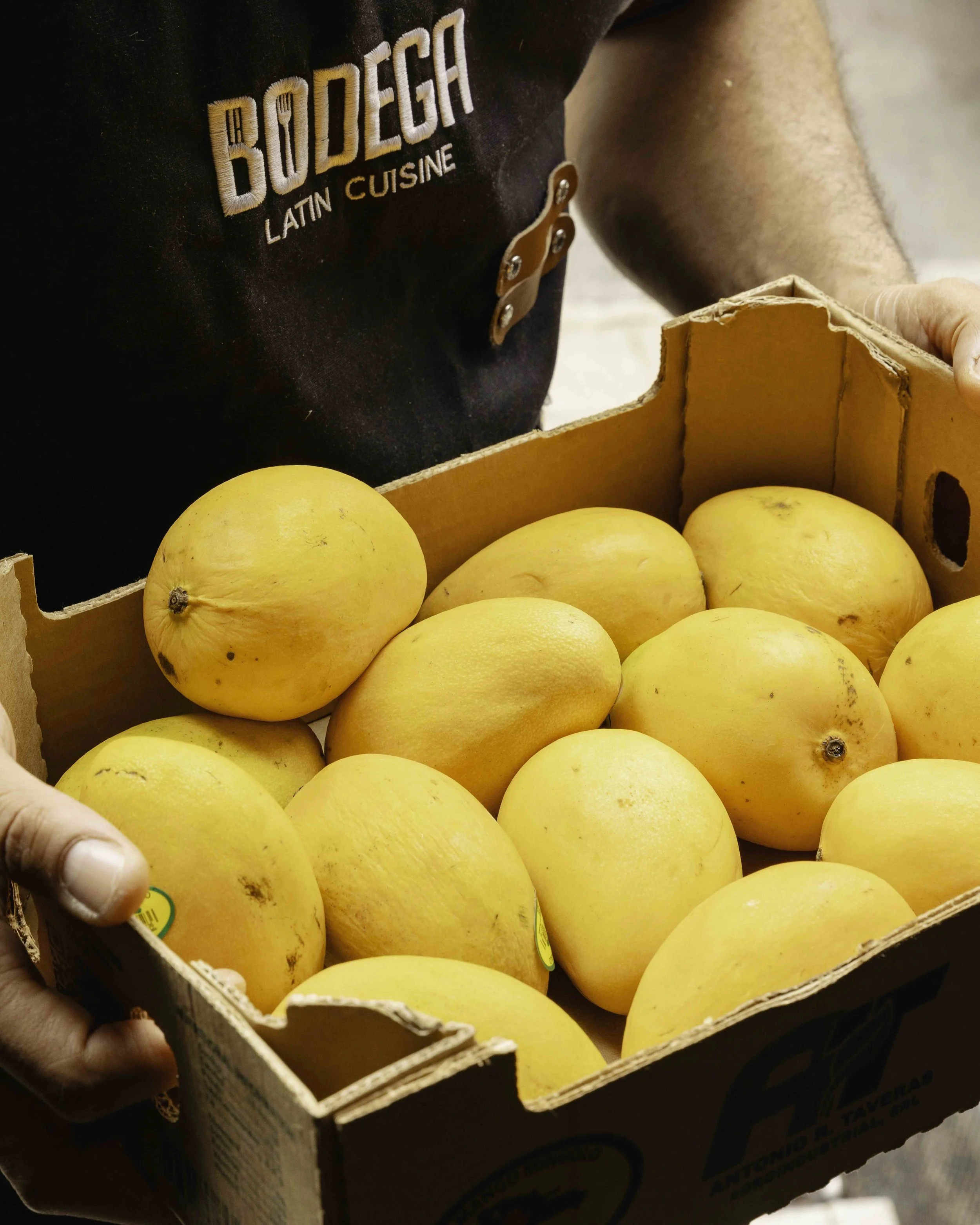 Person holding a cardboard box filled with yellow melons at a market or grocery store.