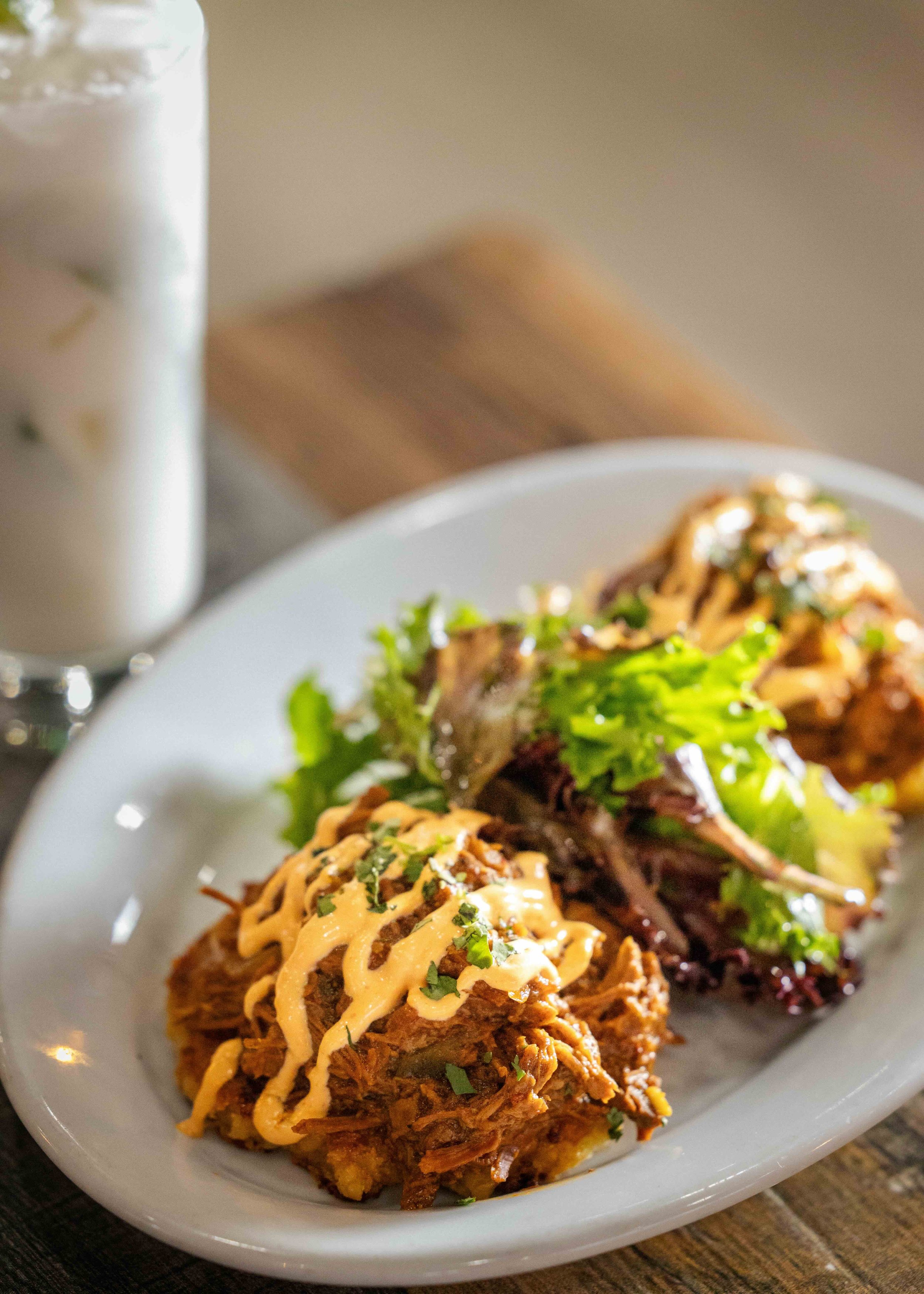 A white plate with pulled pork tacos topped with drizzled sauce and garnished with chopped herbs, served with a side of mixed greens, next to a glass of water on a wooden table.
