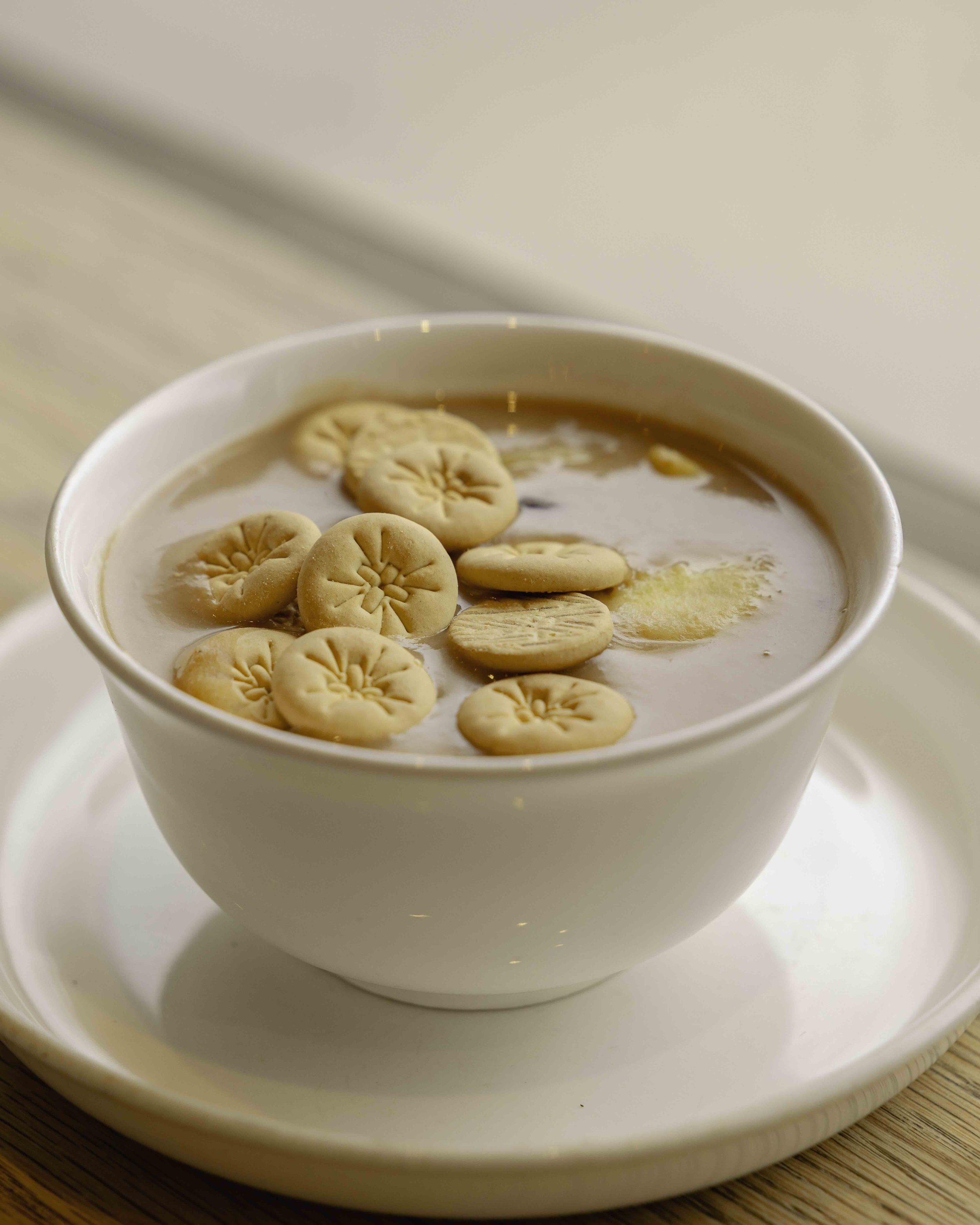 A white bowl of tea with biscuits floating on top, placed on a white saucer on a wooden surface.