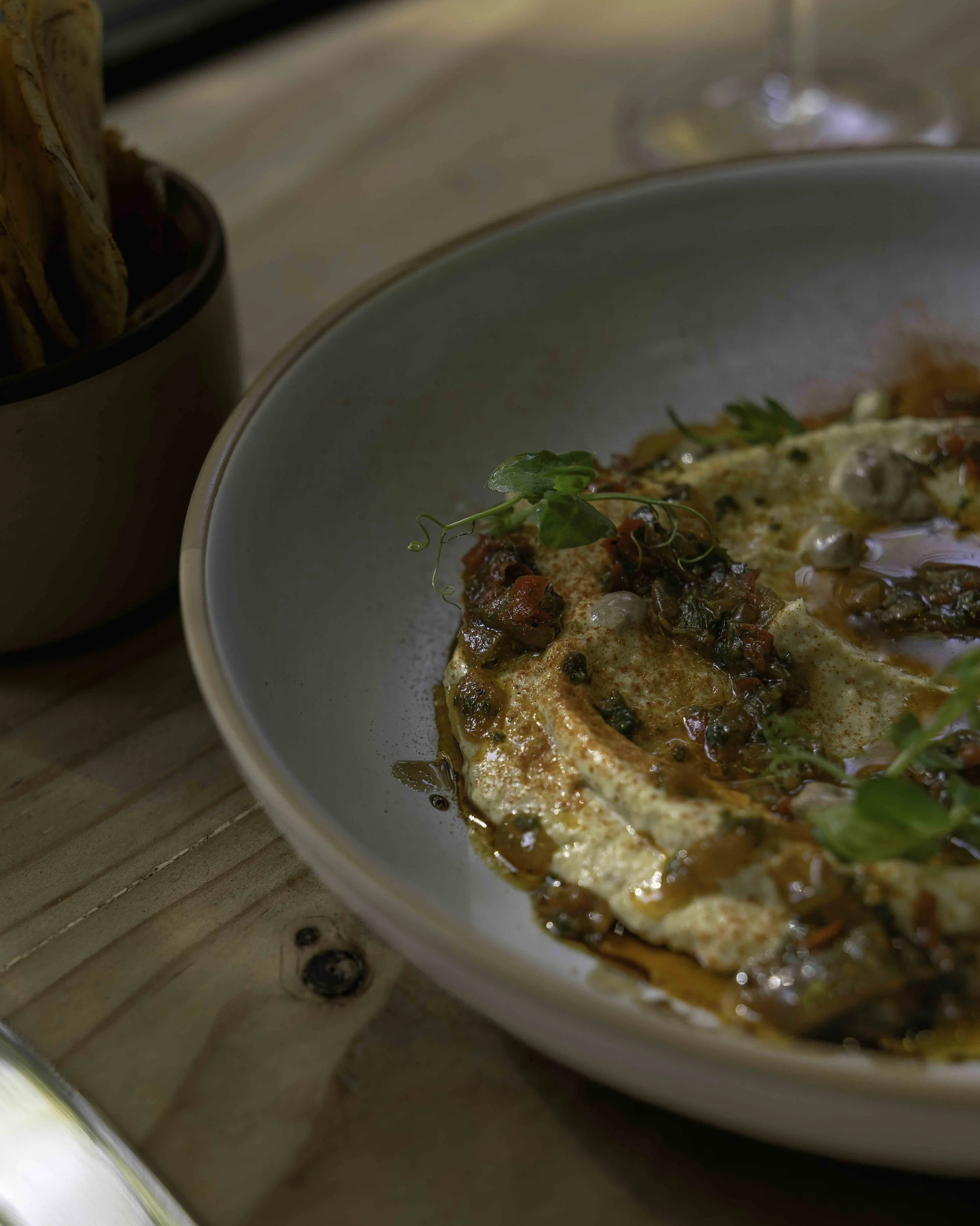 Close-up of a bowl of creamy hummus topped with chopped pico de gallo and garnished with microgreens, with a small dish of breadsticks in the background on a wooden table.