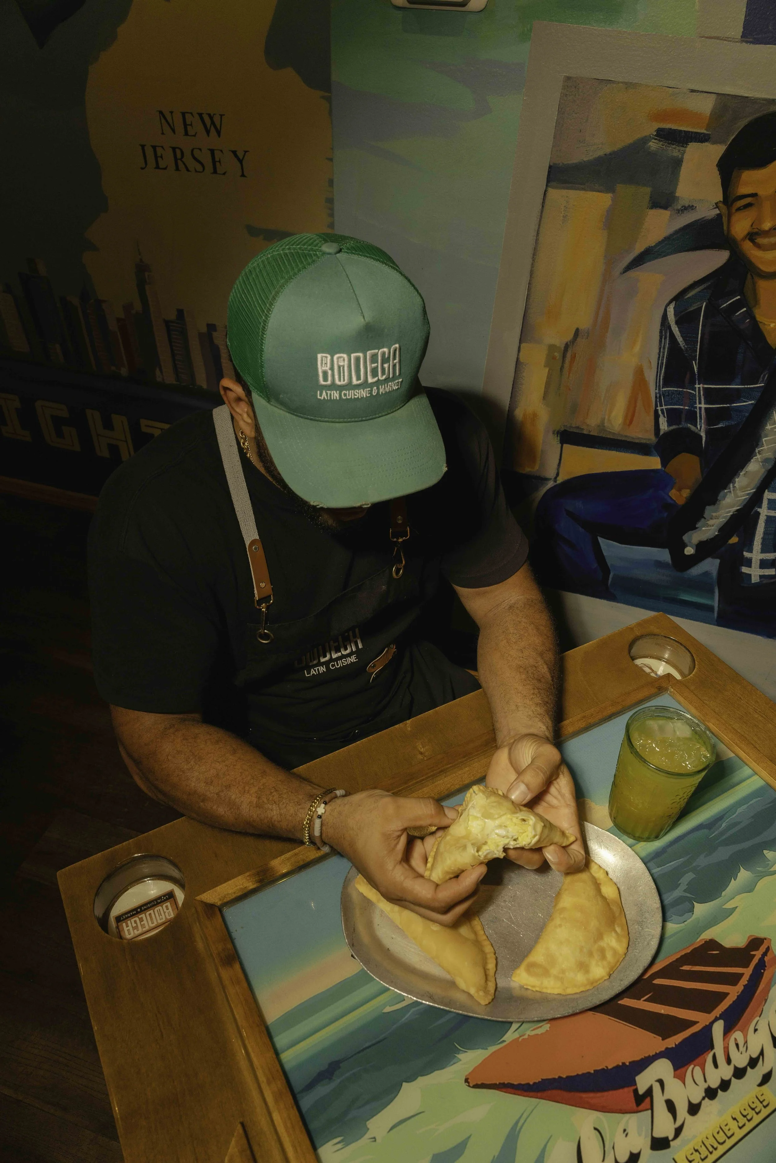 Man wearing a green cap and black apron sitting at a table with a plate of two pasties and two drinks, in front of colorful wall art.