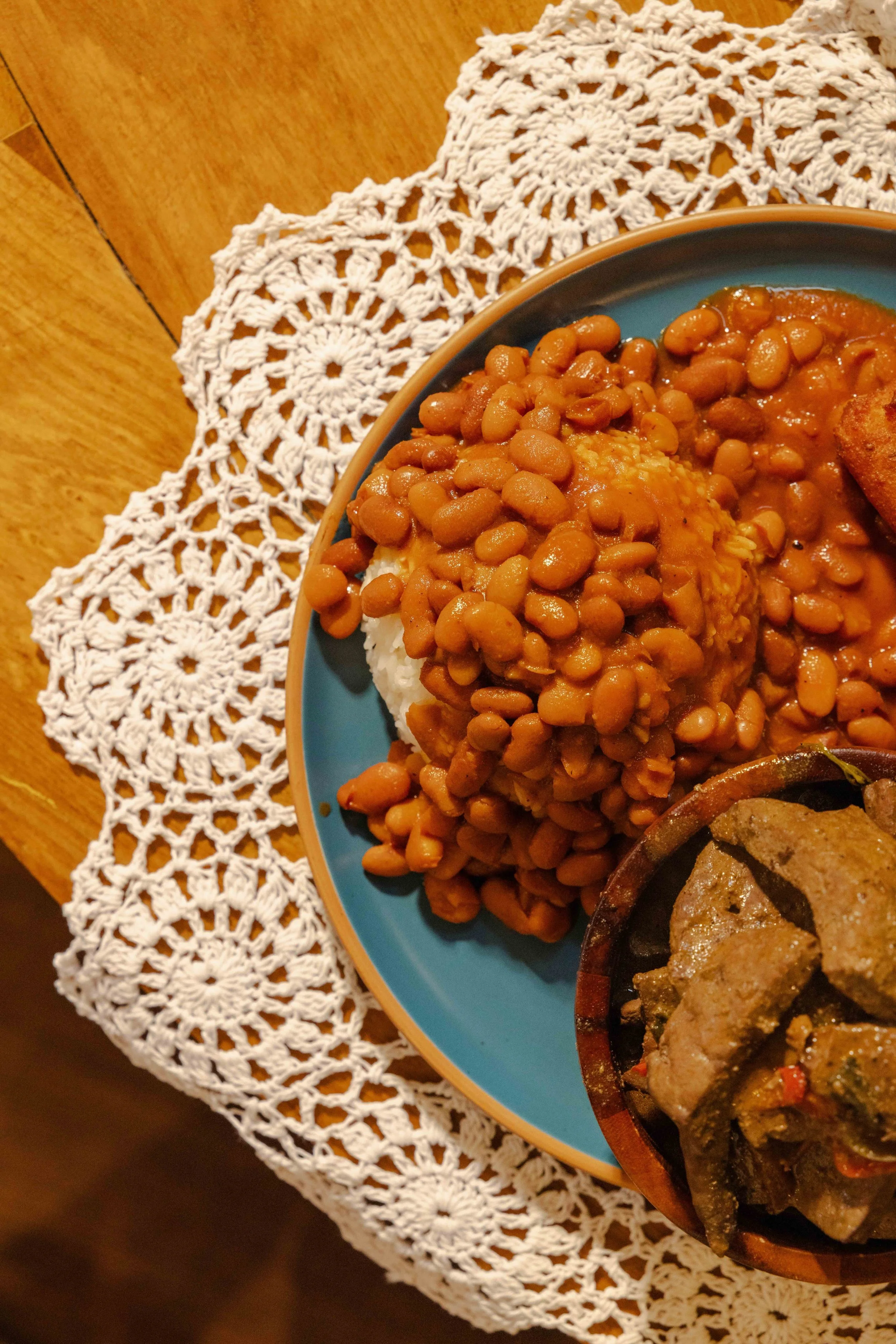 Plate of cooked rice topped with brown beans and a side dish of meat in sauce on a lace tablecloth.