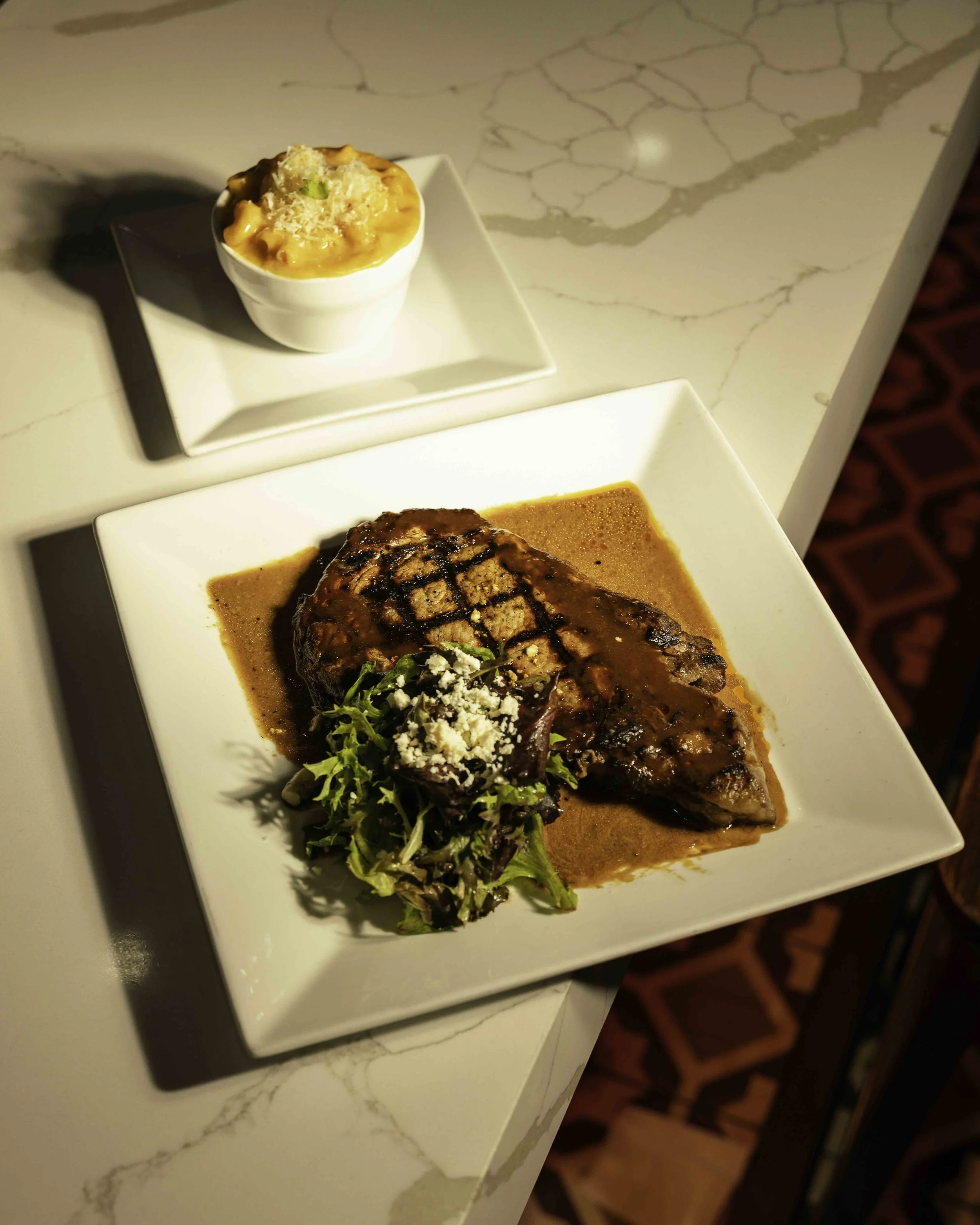 A plate with a cooked steak topped with cheese and greens, served with gravy and a side of salad. A small bowl of macaroni and cheese on a square white plate in the background.