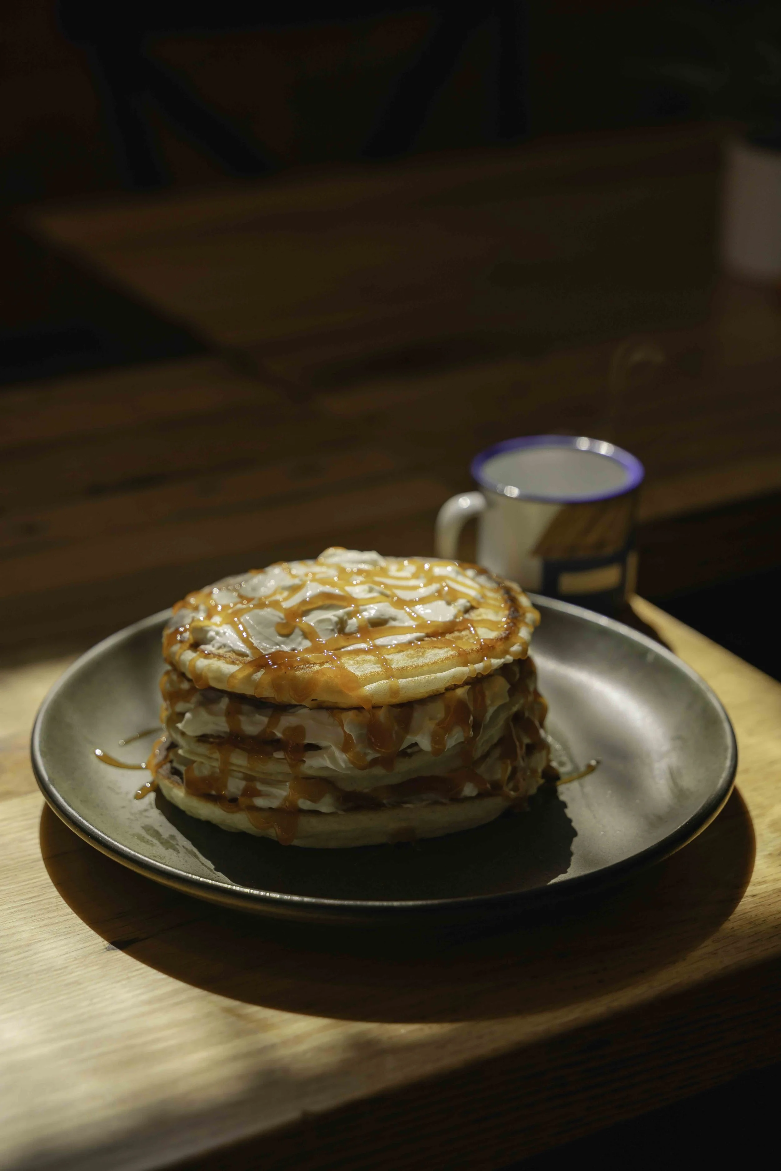 Stack of three pancakes with whipped cream, caramel syrup, on a black plate, with a mug in the background.